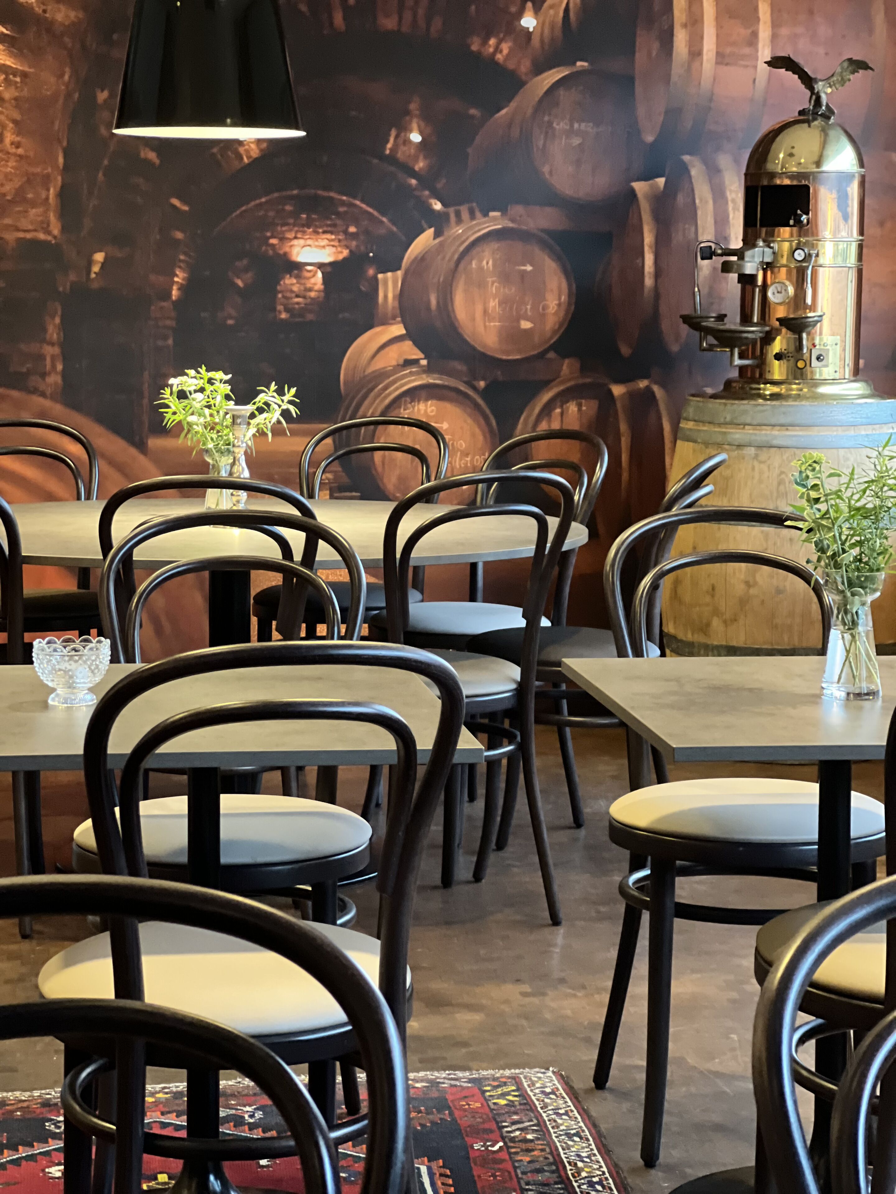 A restaurant interior with multiple tables and chairs, a decorative wall with barrels, and a vintage distillation apparatus.
