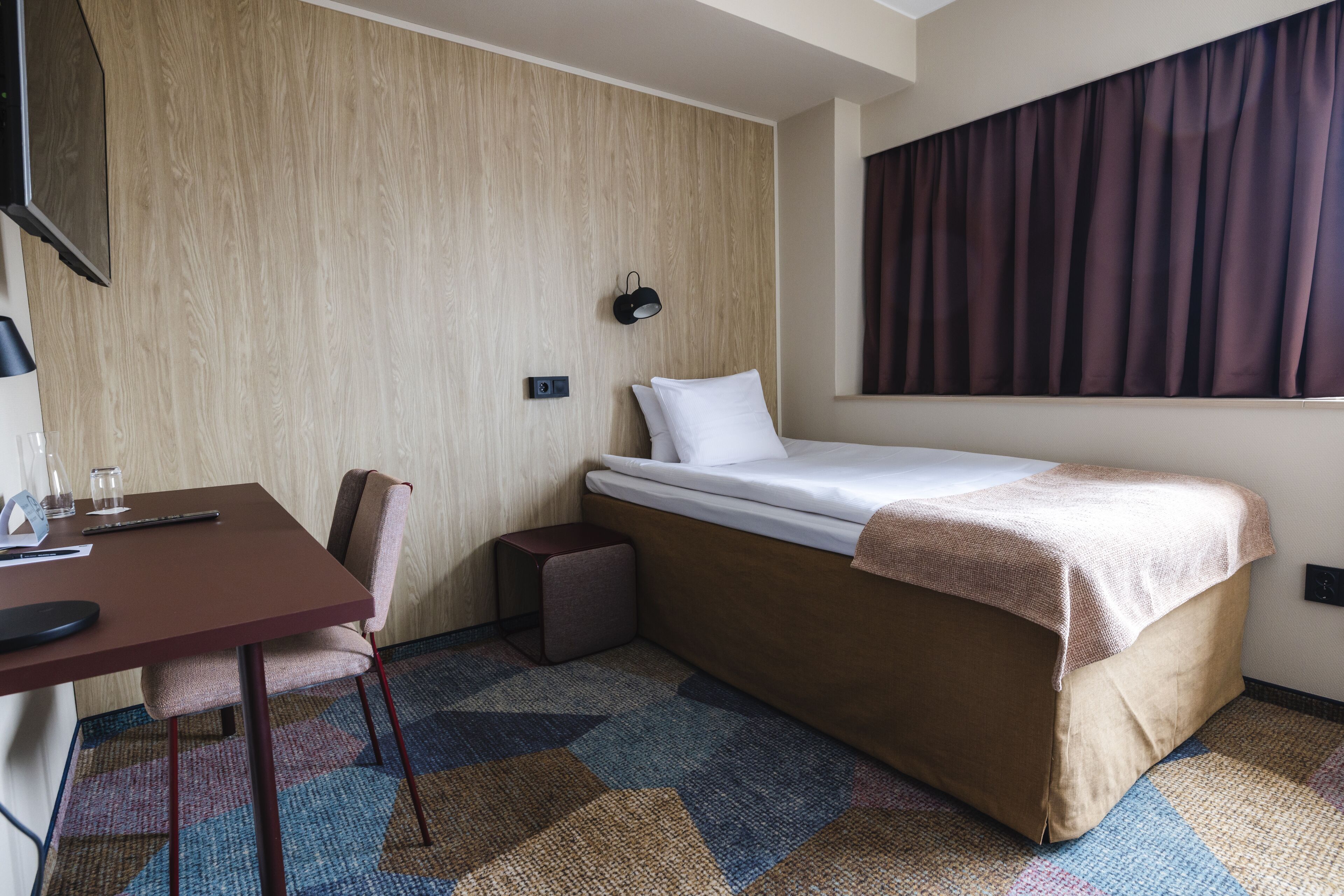A hotel room with a single bed, a desk with a chair, a small table, a wall-mounted TV, and a window with curtains.