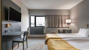 A hotel room with a bed, a desk with a chair, a TV mounted on the wall, a window with curtains, and a lamp on the bedside table. - Quality Hotel Royal Corner (Växjö)