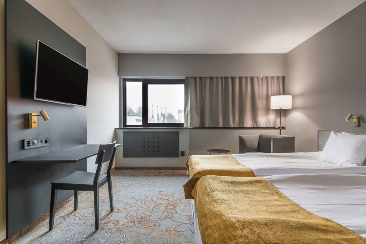 A hotel room with a bed, a desk with a chair, a TV mounted on the wall, a window with curtains, and a lamp on the bedside table.