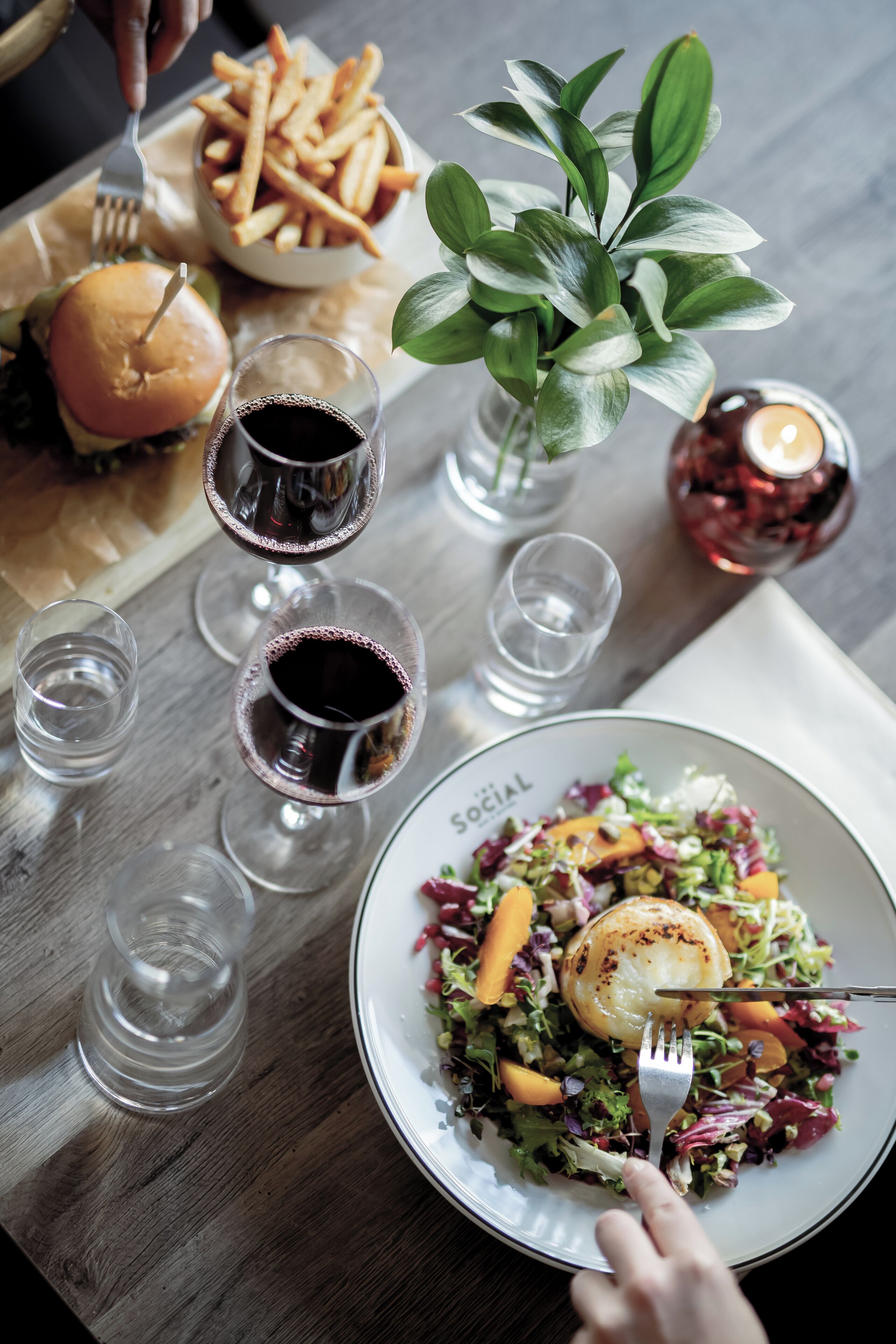 A dining table with a plate of salad, a burger, and a bowl of fries. There are also several glasses of a dark beverage and a lit candle.