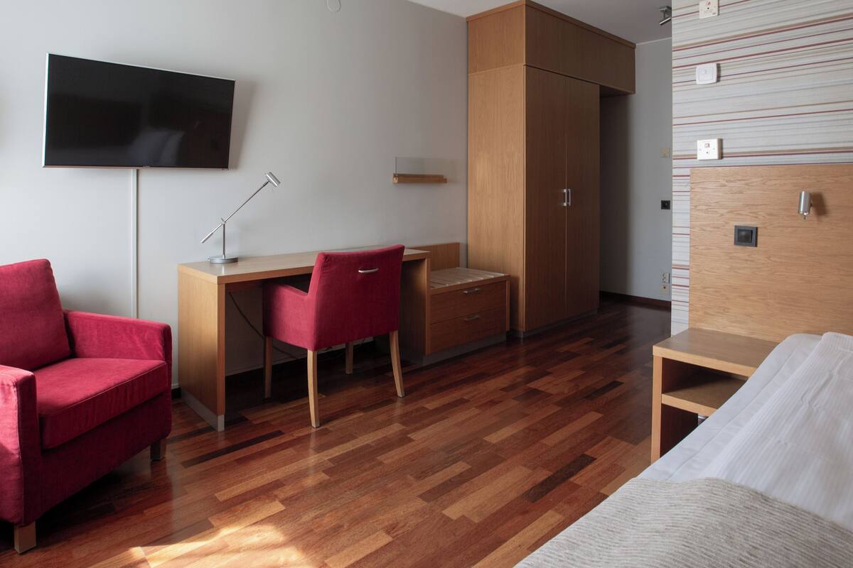 A modern hotel room with a wooden desk, a red chair, a bed, and a TV mounted on the wall.