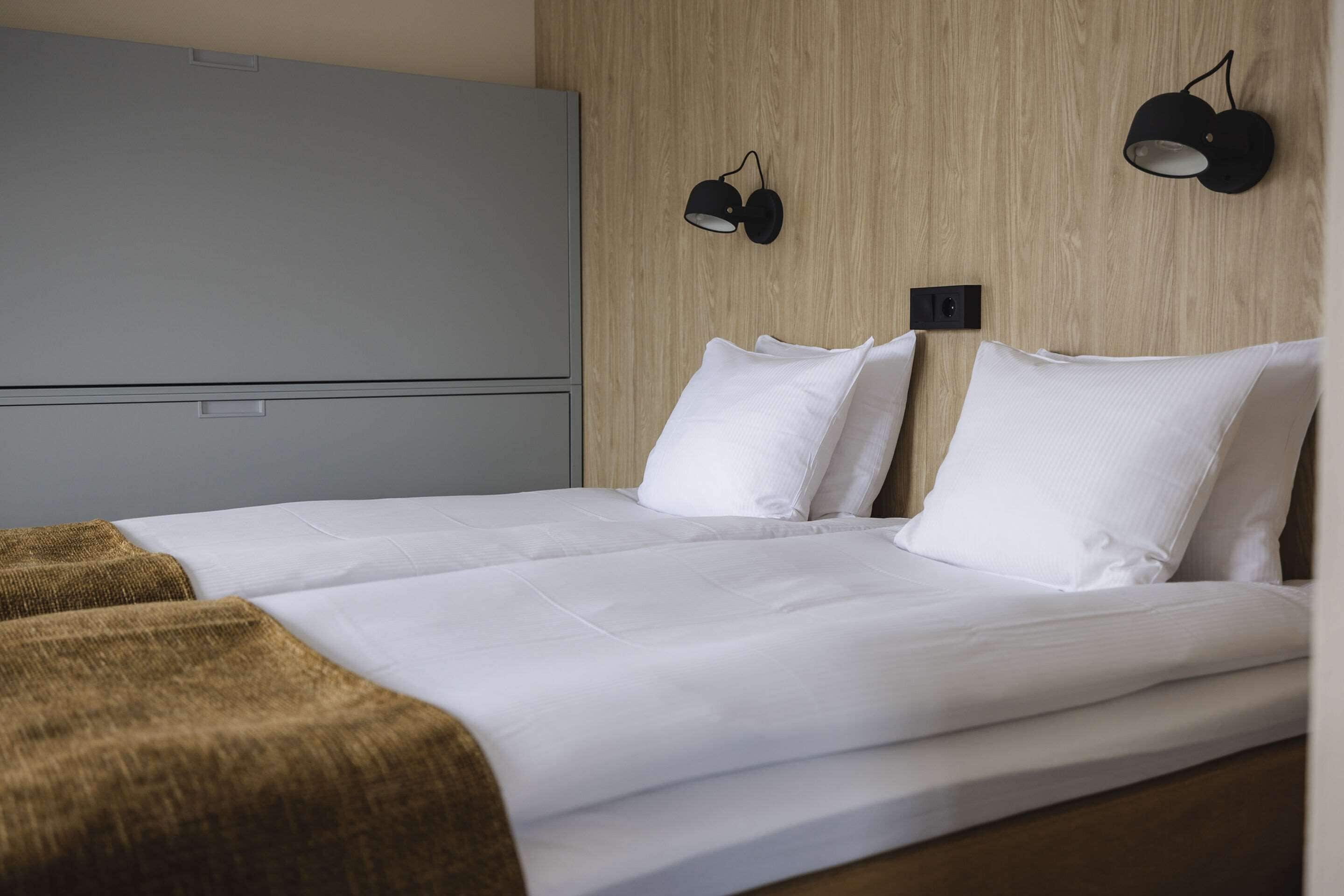 A neatly made bed with white linens and two pillows, a grey wardrobe, and two wall-mounted lamps.