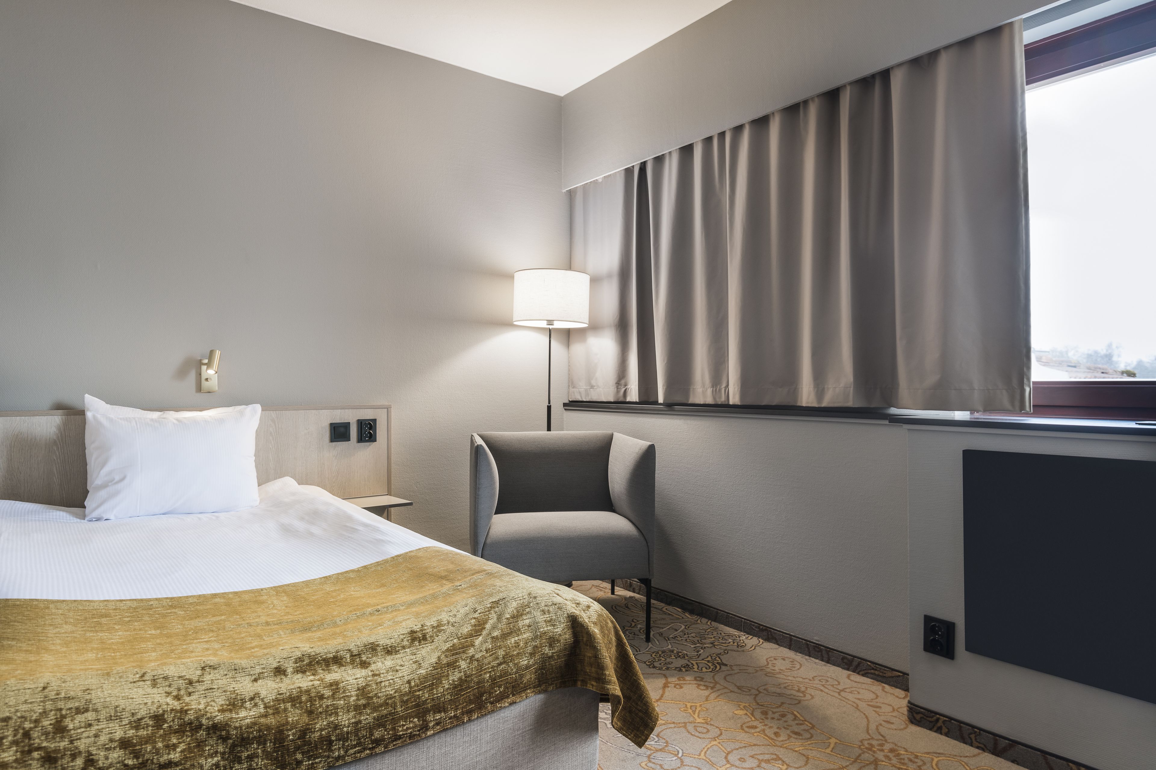 A hotel room with a bed, a chair, a lamp, and a window with curtains.