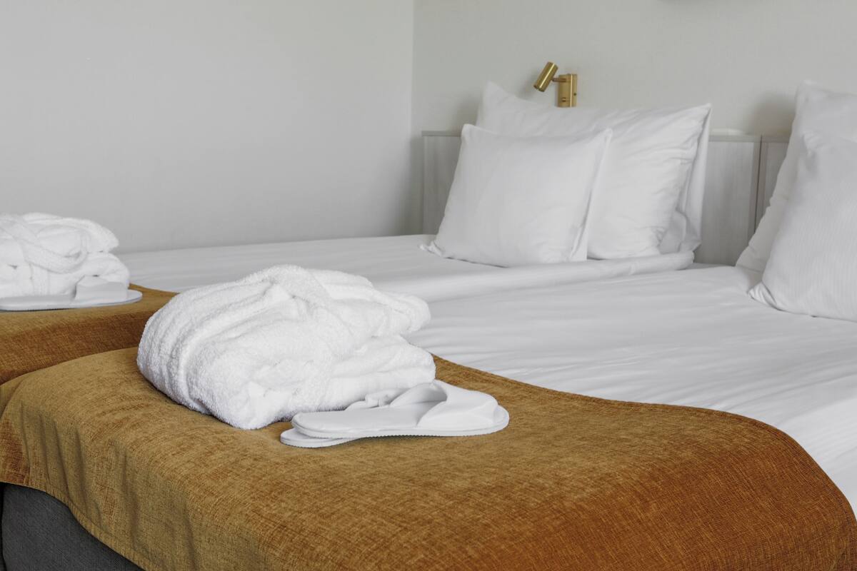 Two beds with white linens, a brown throw blanket, and a white bathrobe on the left bed.