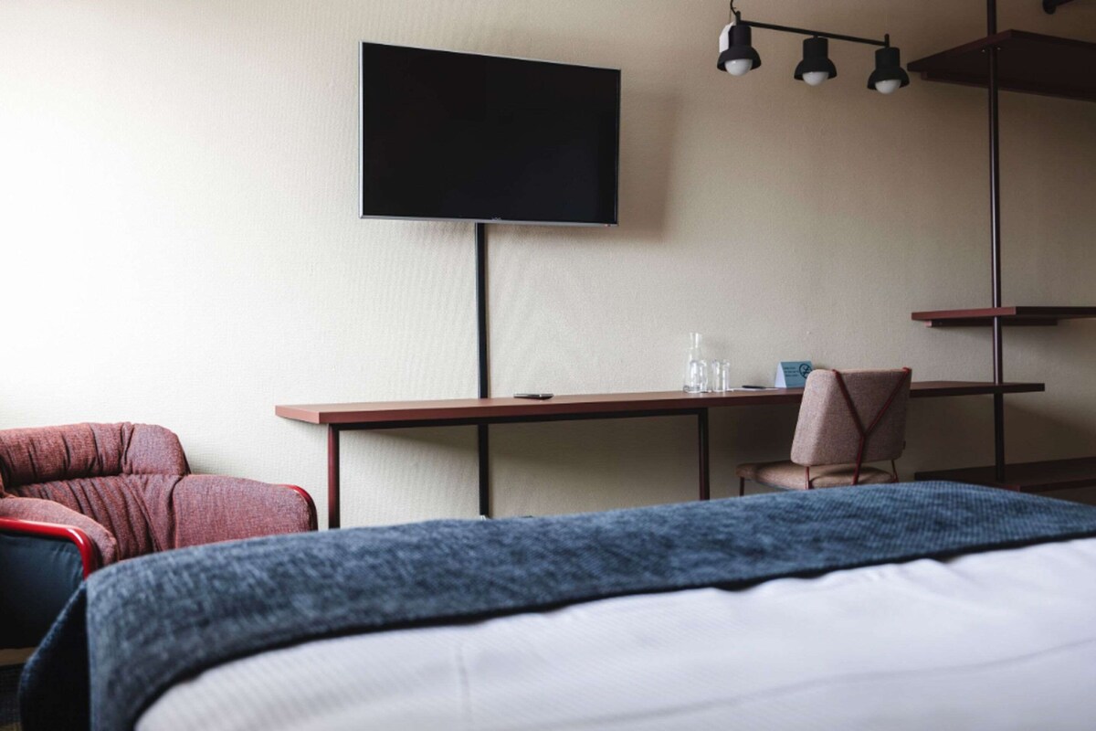 A hotel room with a bed, a desk, a chair, a television, and a small table with a glass and a bottle.