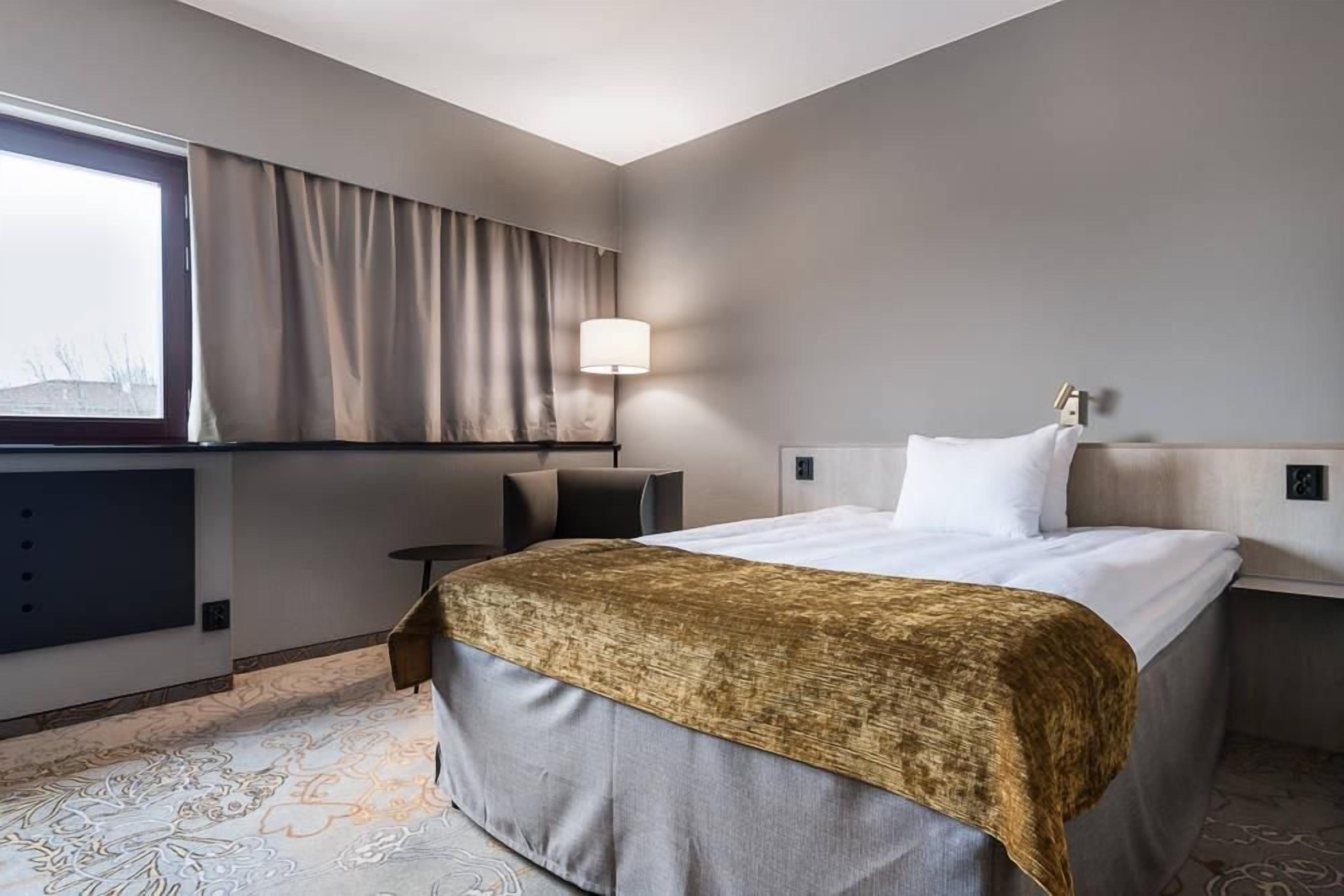 A hotel room with a bed, a desk, a chair, a lamp, and a window with curtains.