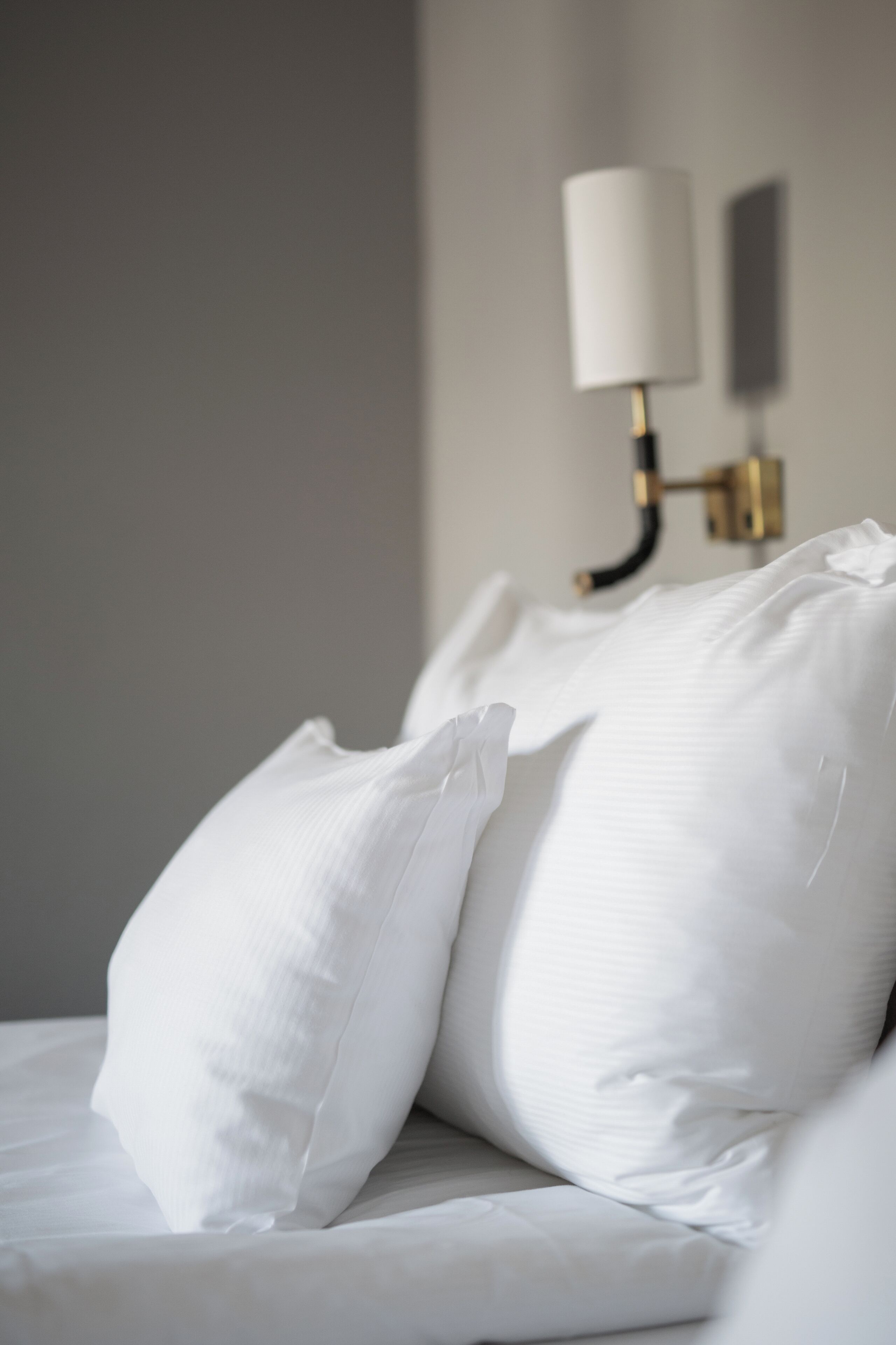 Ein ordentlich bezogenes Bett mit weißen Leinen und Kissen, eine Wandlampe und ein schlichtes, aufgeräumtes Zimmerdesign.