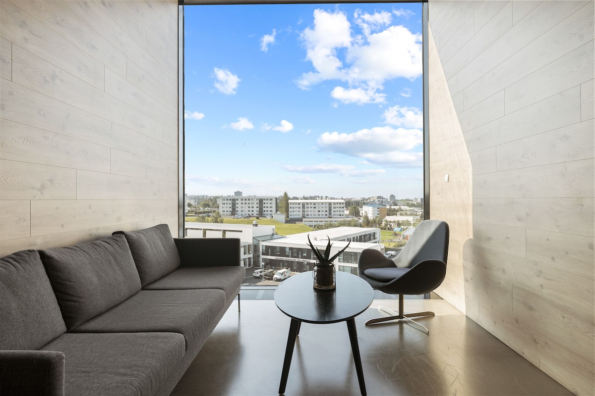 Ein modernes Wohnzimmer mit einem grauen Sofa, einem schwarzen Sessel und einem kleinen Tisch mit einer Pflanze. Ein großes Fenster bietet Blick auf Gebäude und einen klaren Himmel.