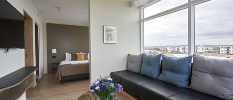 A modern hotel room with a large window, a sofa, a bed, and a flat-screen TV.