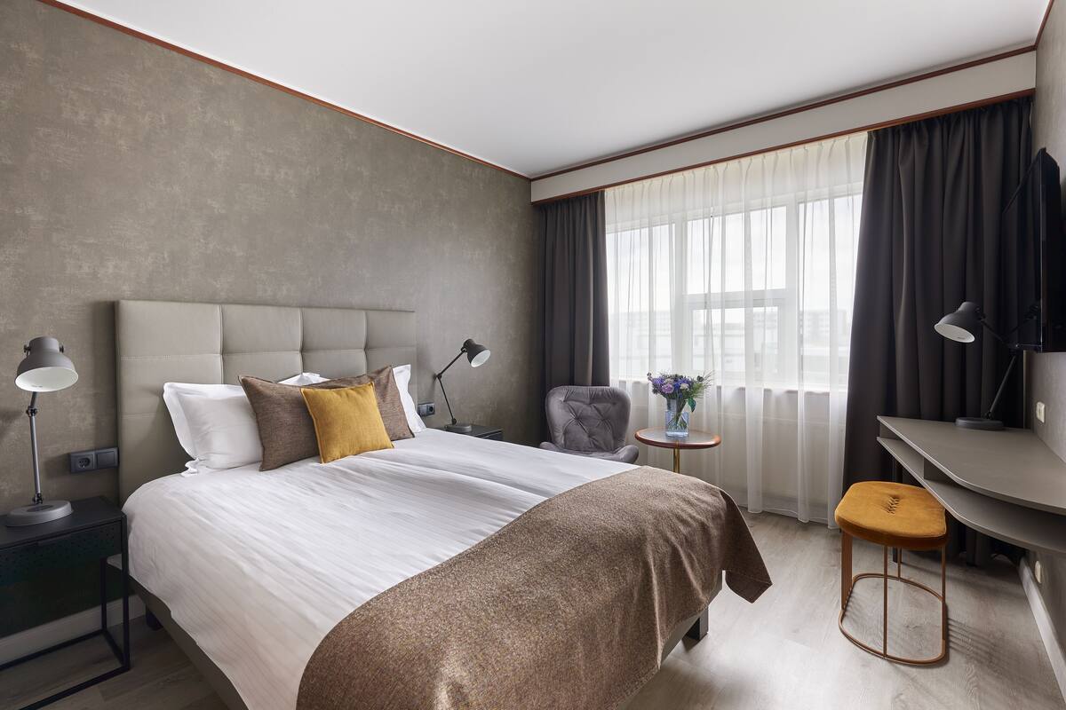 Ein modernes Hotelzimmer mit einem großen Bett, einem Schreibtisch, einem Sessel und einem Fenster mit Vorhängen.