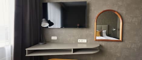 Una habitación moderna con un televisor montado en una pared gris, un espejo con marco de madera, una mesa redonda pequeña y un taburete amarillo.