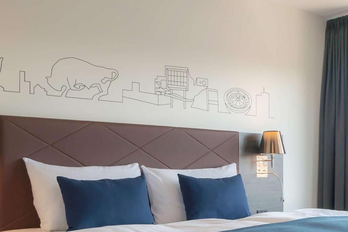 A hotel room with a bed, a headboard, a wall art depicting a cityscape, a window with a view of a cruise ship, and a lamp.