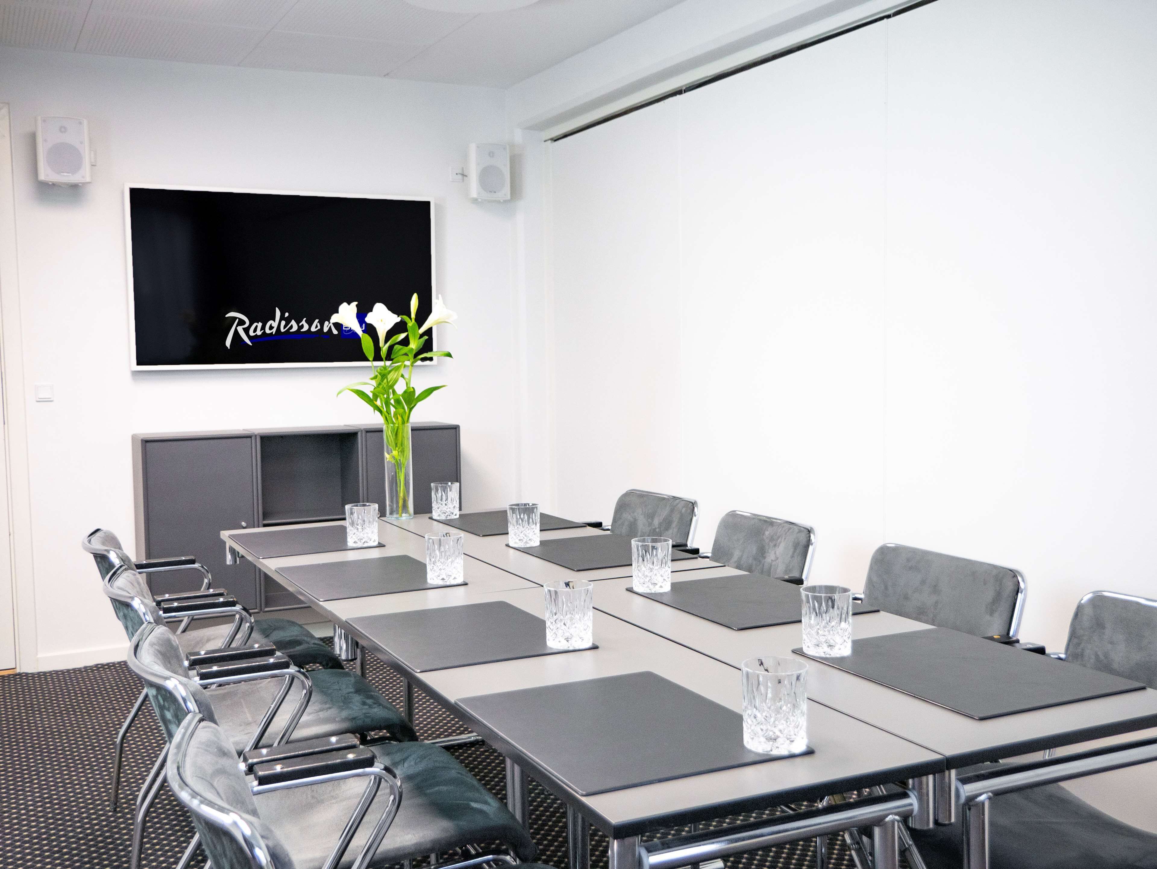 A conference room with a long table, grey chairs, and a TV mounted on the wall.