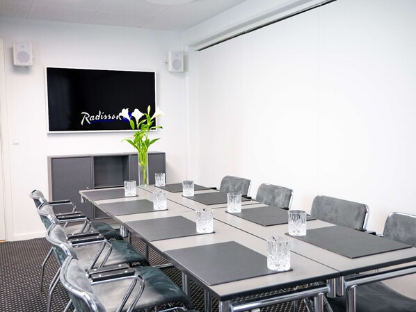 A conference room with a long table, grey chairs, and a TV mounted on the wall.