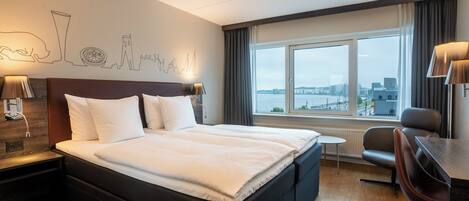 A hotel room with a large bed, a desk, a chair, a view of the city, and a wall with a cityscape mural.