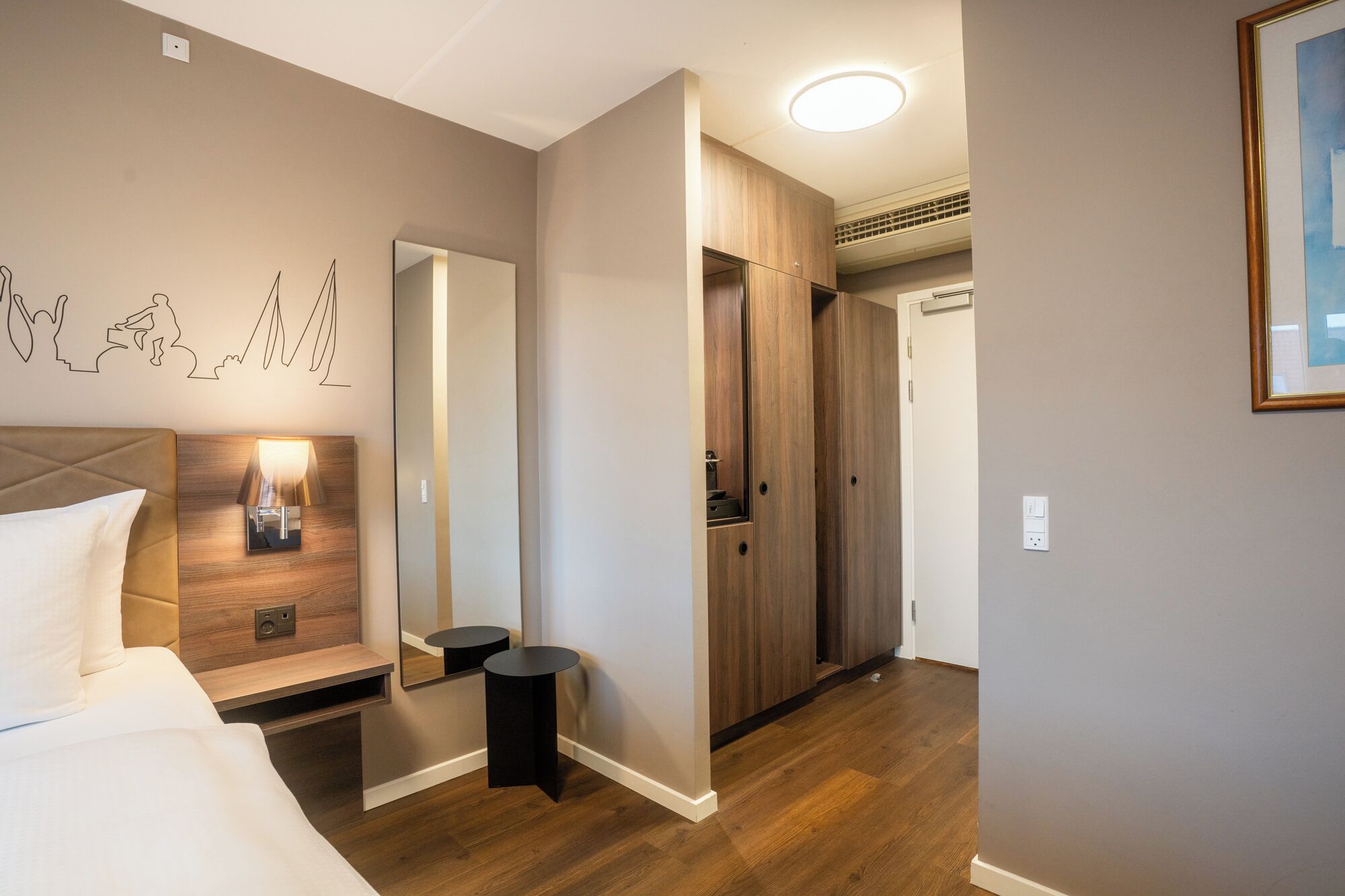 Habitación de hotel con cama, mesita de noche, espejo, armario y un pequeño taburete.