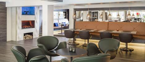 A modern interior with a dining area featuring round tables and green chairs, a bar area with pendant lighting, and a visible fireplace.
