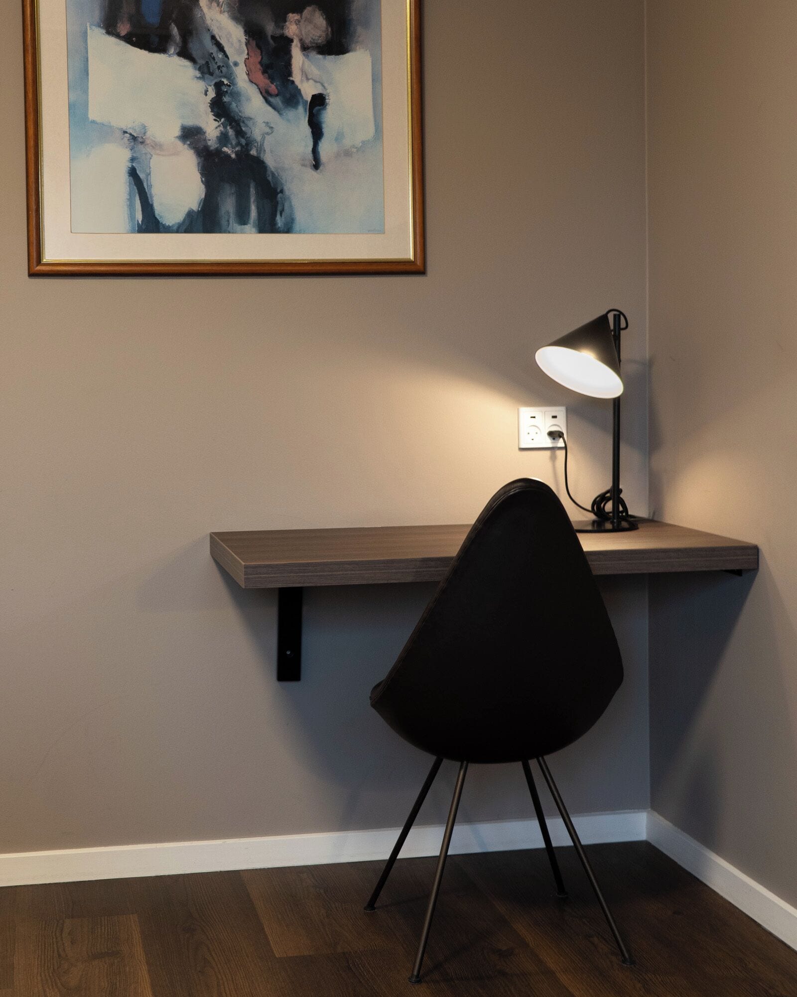 A corner room with a desk, chair, lamp, and a painting on the wall.