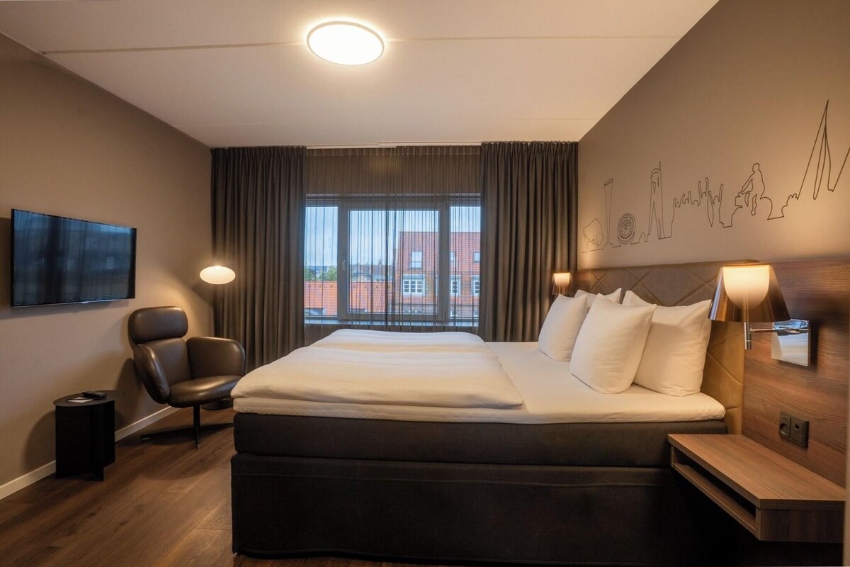 A hotel room with a large bed, a chair, a TV, and a view of buildings through the window.