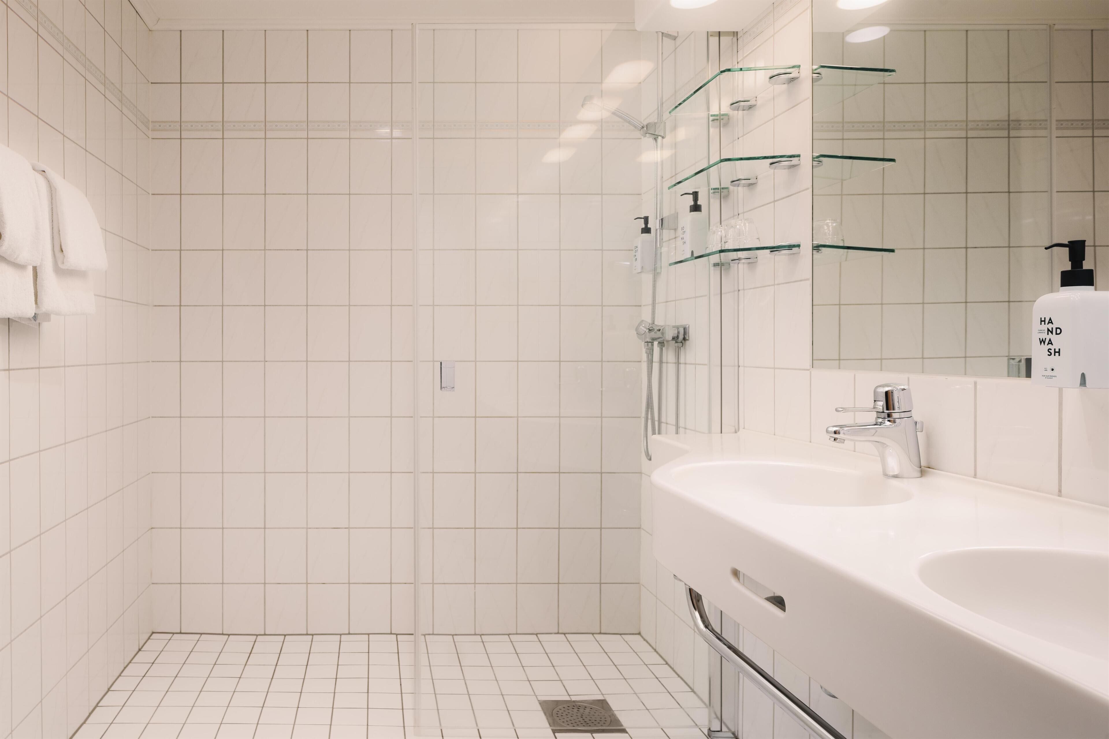 Un baño moderno con azulejos blancos, cabina de ducha de vidrio y un espejo grande con dispensador de gel hidroalcohólico.