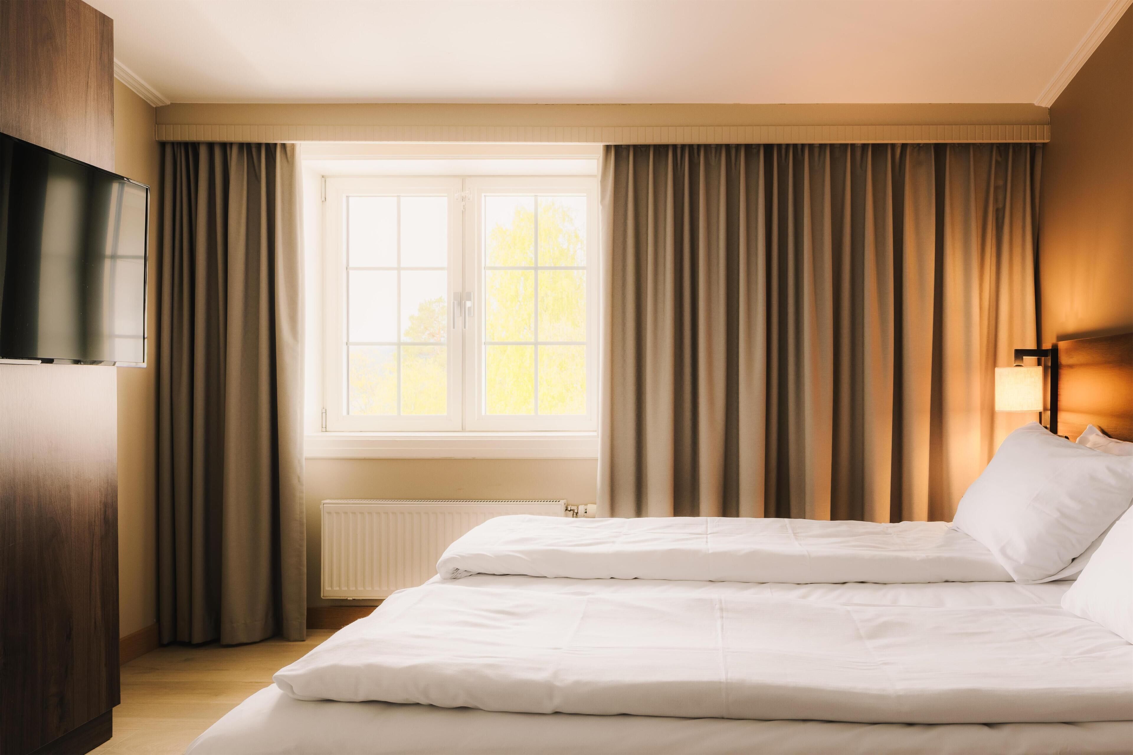A hotel room with a bed, a TV, a window with curtains, and a radiator.