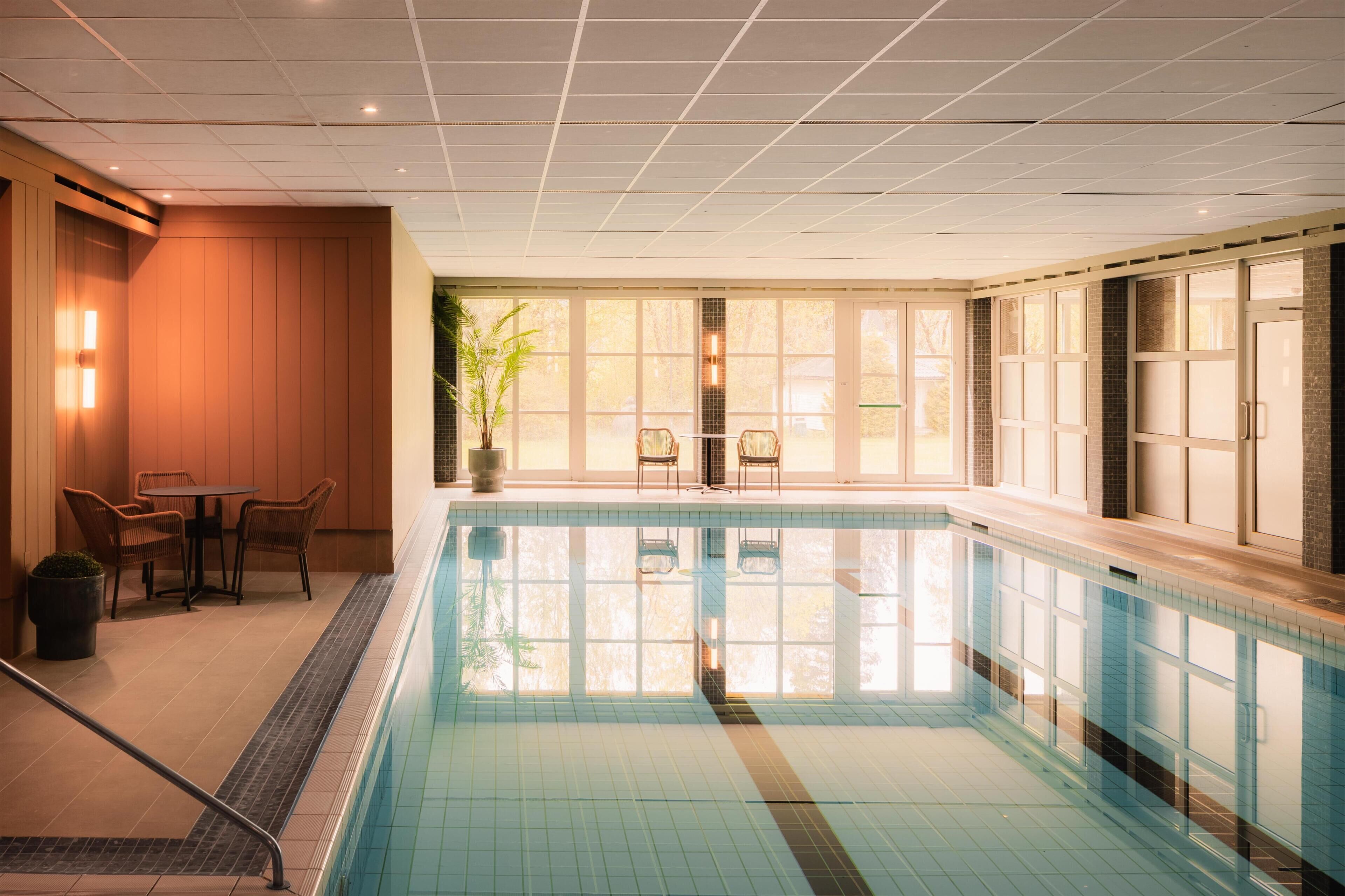 En indendørs swimmingpool med flisegulv, et opholdsområde med stole og store vinduer der lukker naturligt lys ind.
