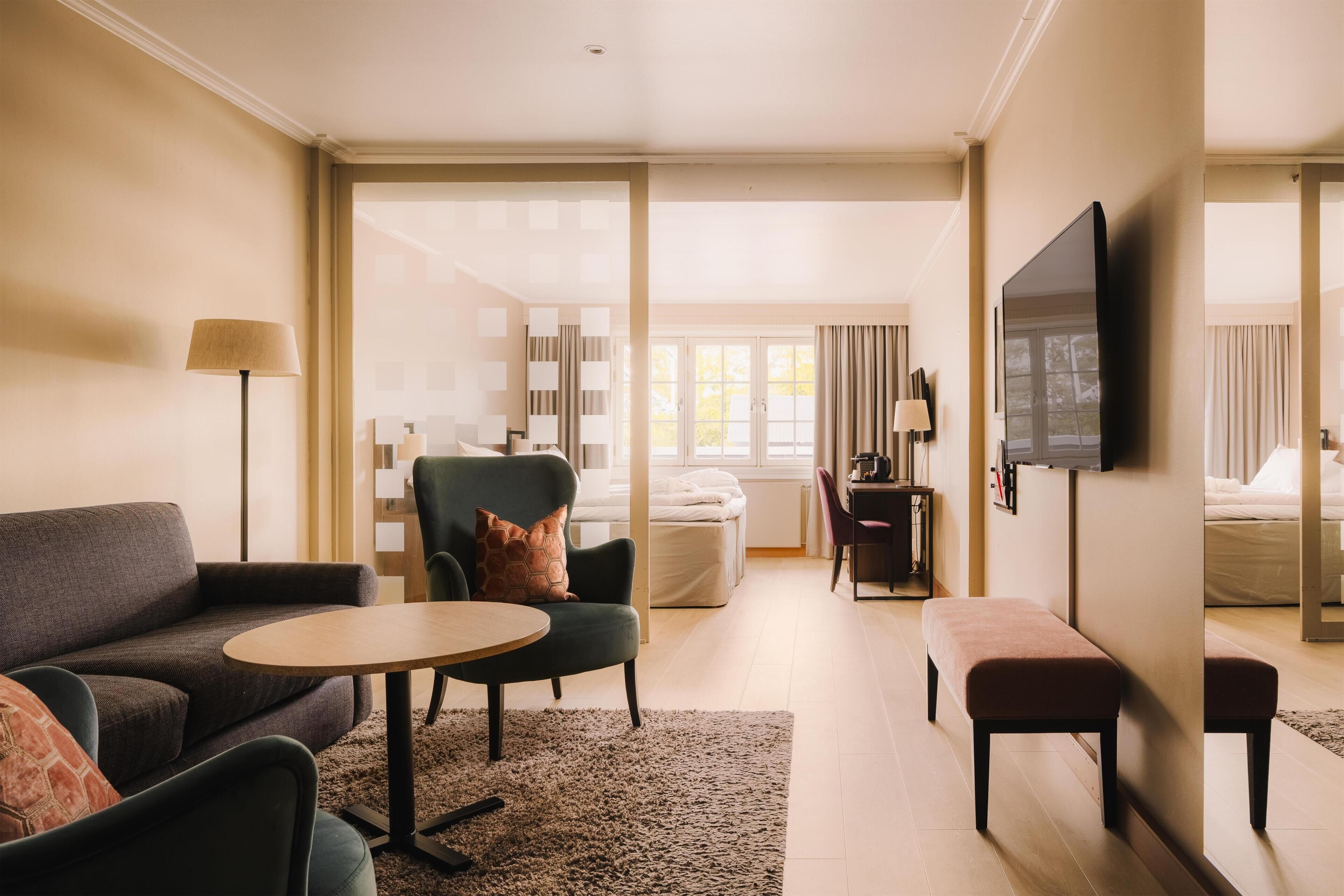 A hotel room with a sofa, armchair, round table, and a TV mounted on the wall.