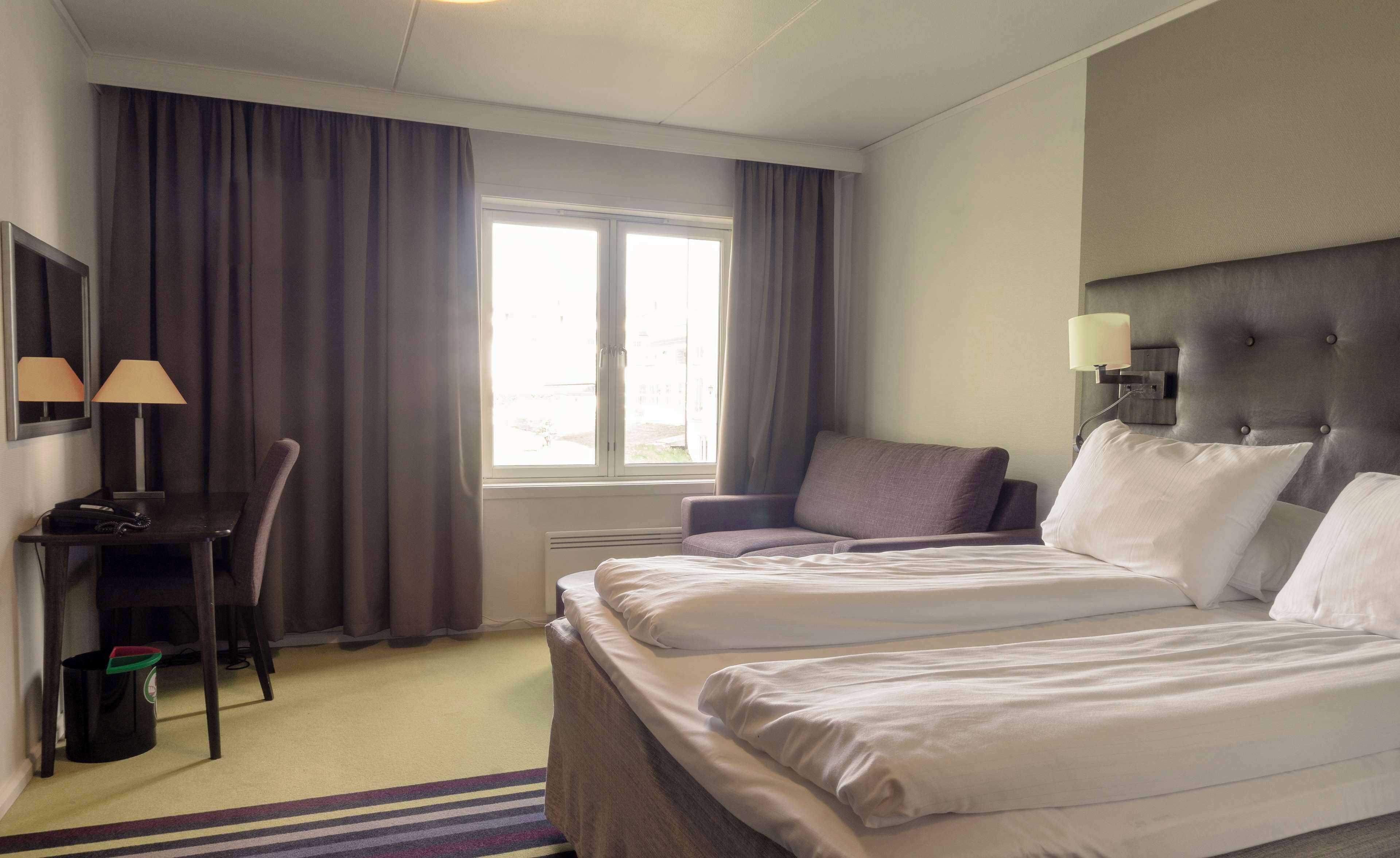 A hotel room with two beds, a desk with a chair, a window with curtains, and a wall-mounted lamp.
