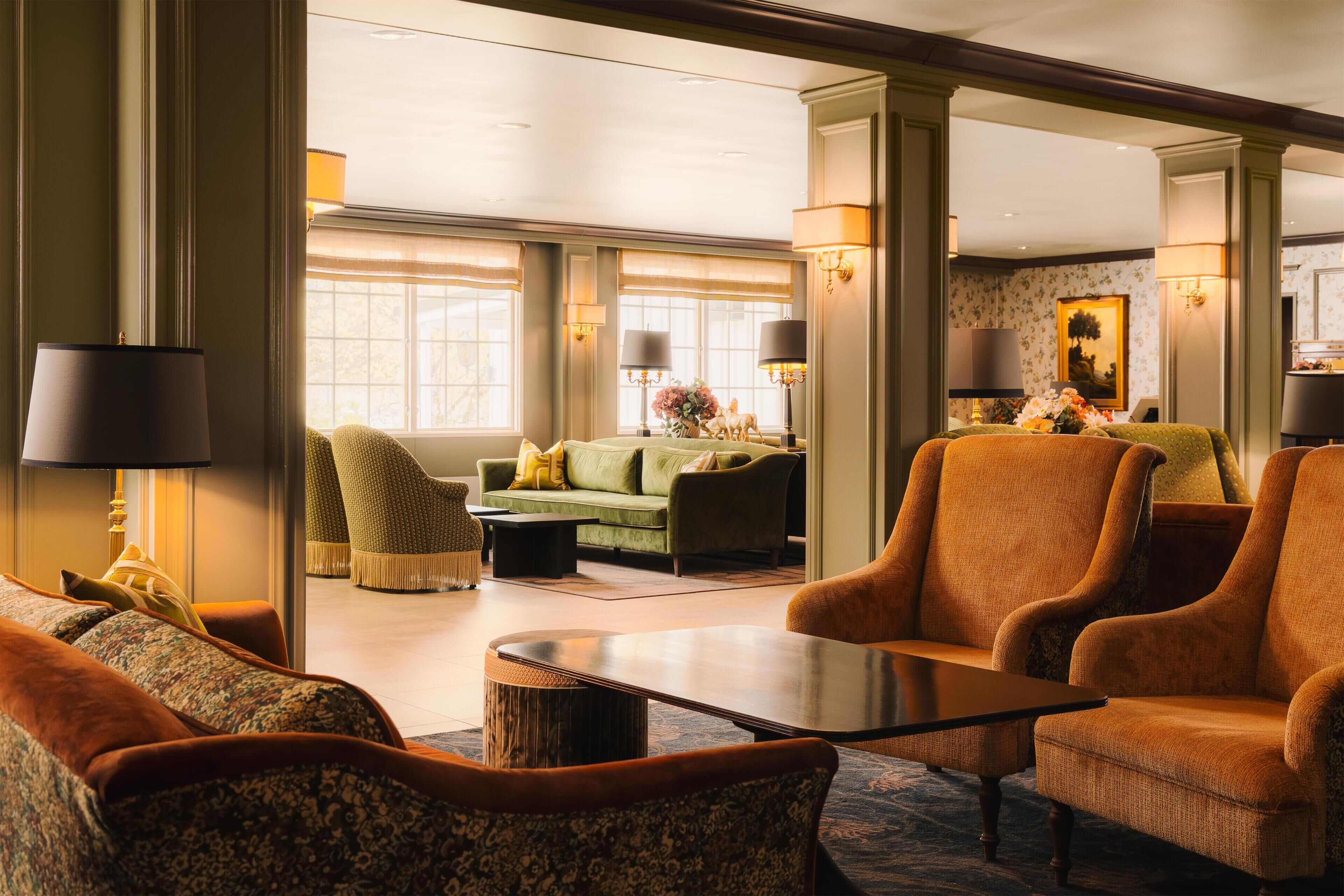 En rymlig hotellobby med en grön soffa, orange fåtöljer, ett soffbord och ett stort fönster som släpper in naturligt ljus.