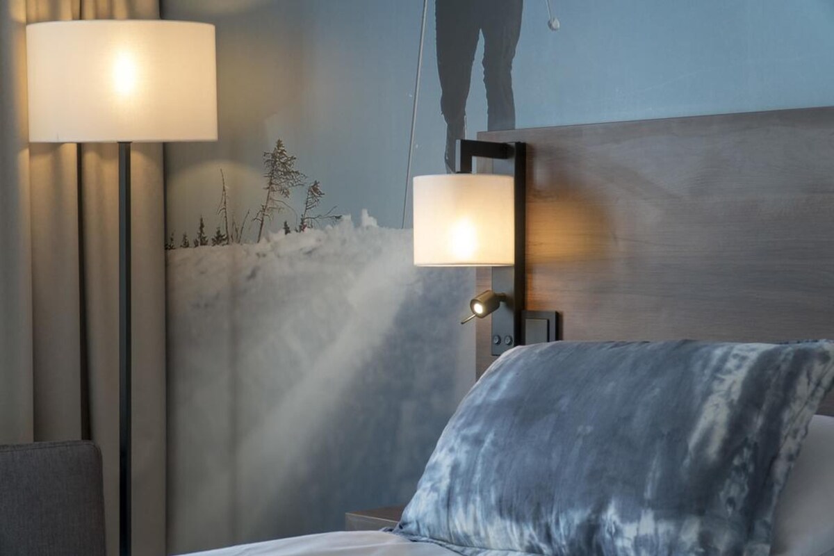 A hotel room with a bed, a nightstand, a lamp, and a wall-mounted light fixture.