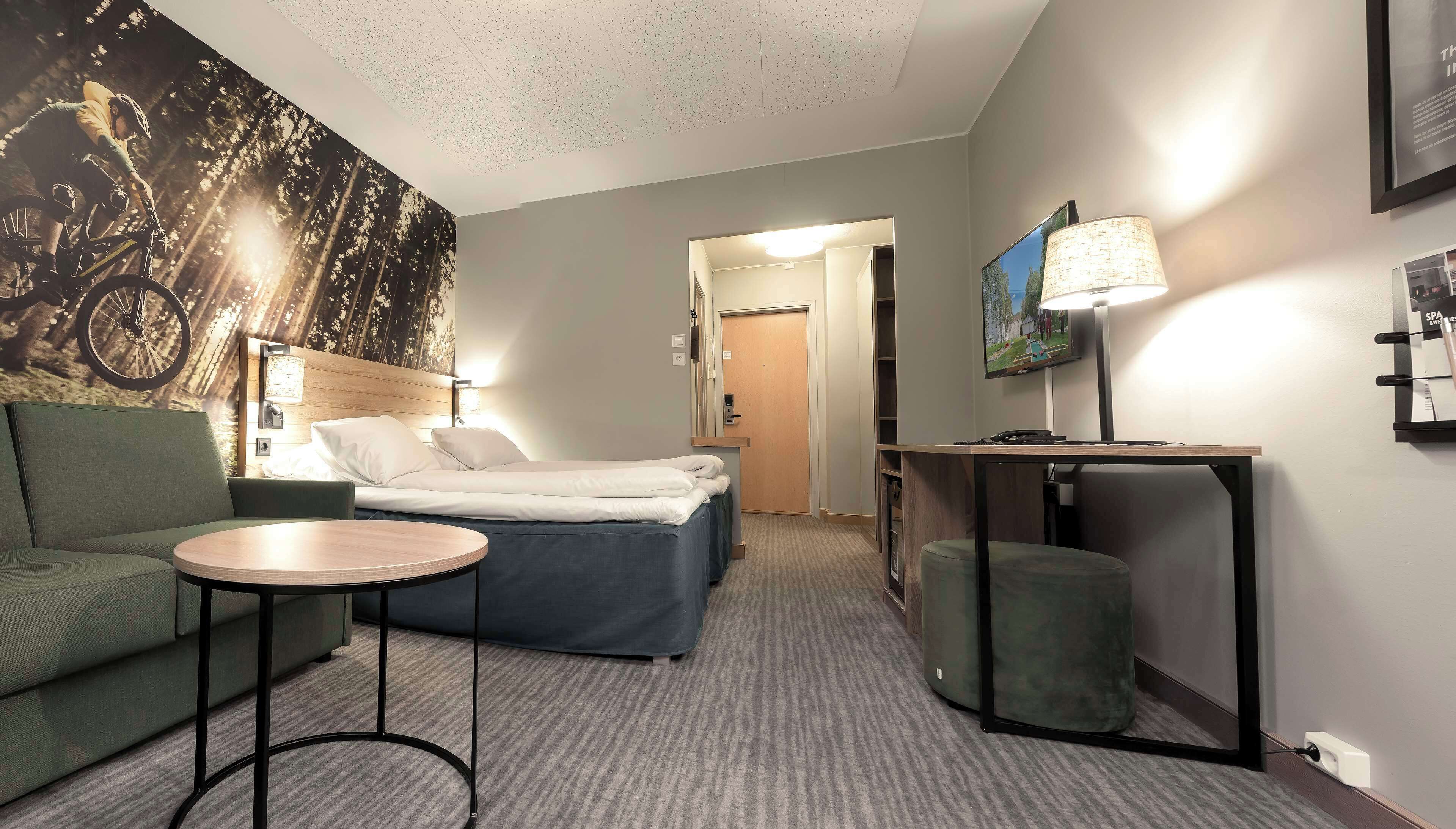 Habitación de hotel con cama, sofá verde, una pequeña mesa redonda, lámpara, televisor y pared con un mural de bicicleta.