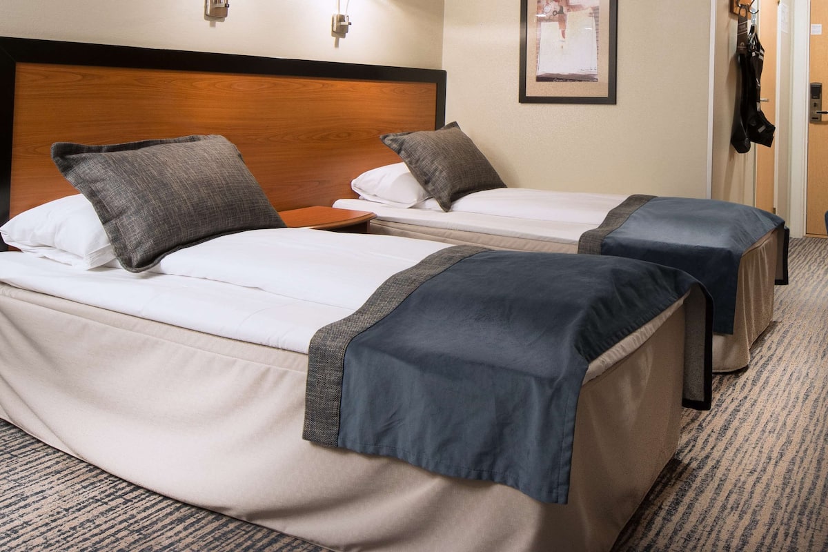 Quarto de hotel com duas camas de solteiro, cabeceira de madeira, uma mesa pequena e uma porta.