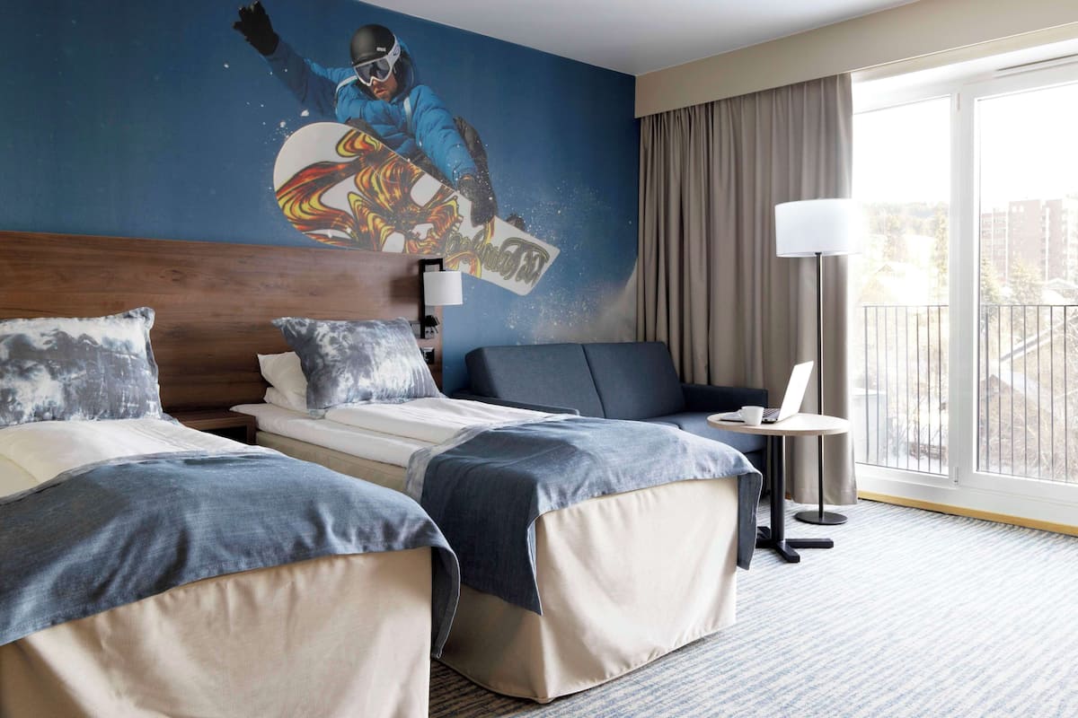 Quarto de hotel com duas camas, uma escrivaninha, um computador portátil, uma luminária e uma janela grande com vista.
