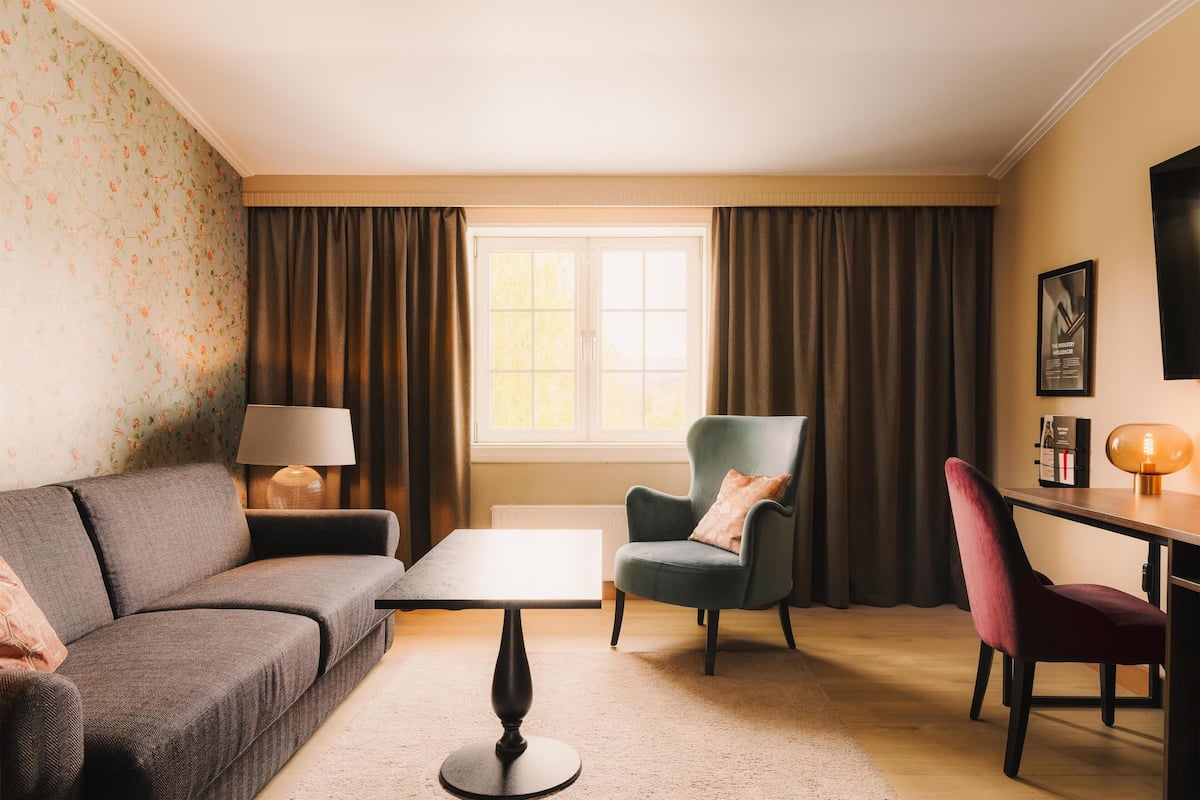 Uma sala de estar com sofá, poltrona, mesa, luminária e uma janela com cortinas.
