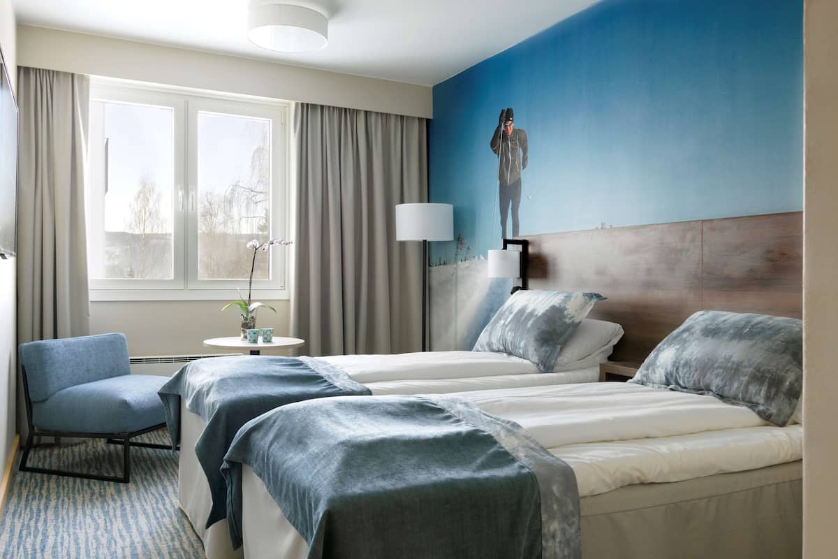 Quarto de hotel com duas camas, uma cadeira, uma mesinha com uma planta e vista para o exterior através de uma janela.