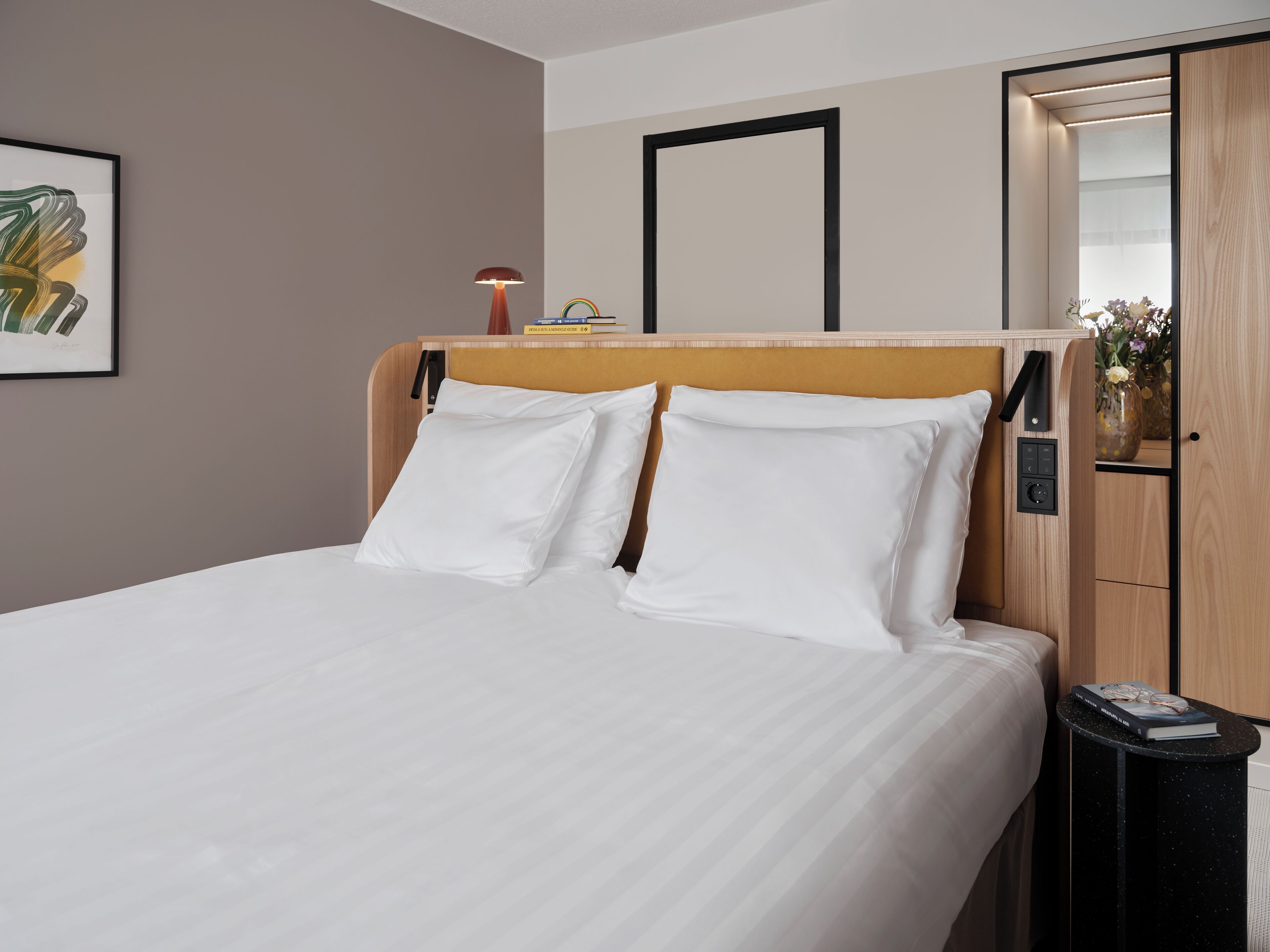 A neatly made bed with white linens, a wooden headboard, and a bedside table with a book and glasses.