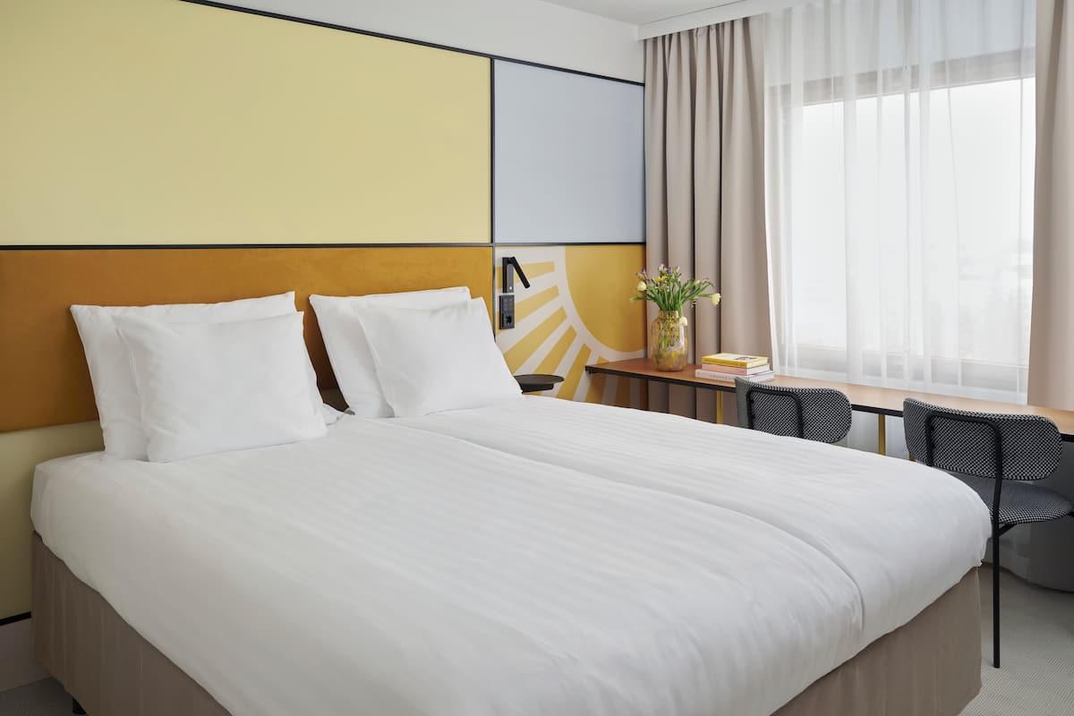 Une chambre d’hôtel avec un grand lit, un bureau avec une chaise, un vase de fleurs et une fenêtre avec des rideaux.