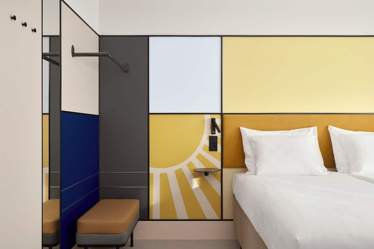 Une chambre d’hôtel moderne, équipée d’un lit, d’une banquette, d’un miroir et d’un mur orné de motifs géométriques.