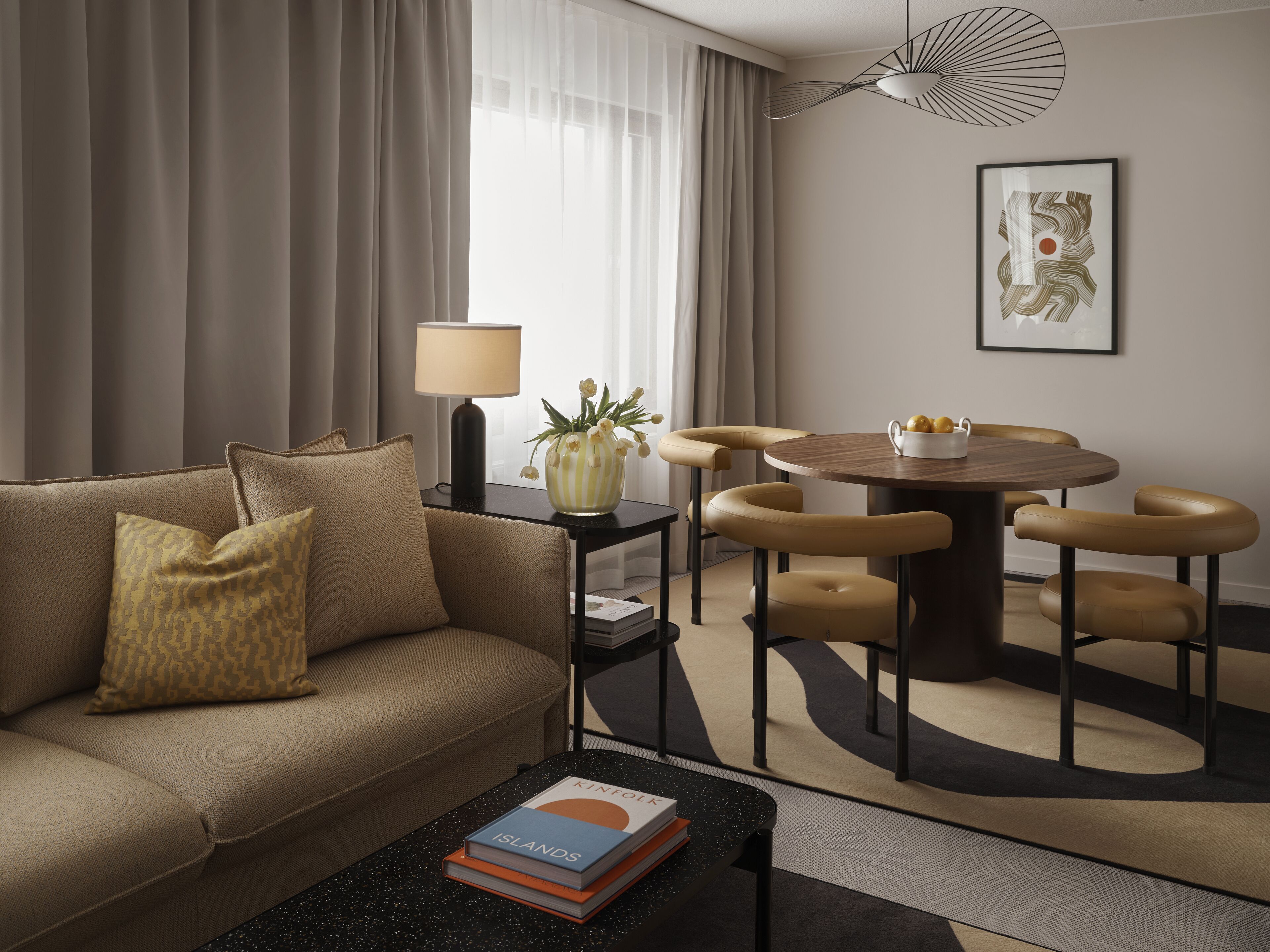 A modern hotel room with a sofa, a coffee table with books, a lamp, a vase with flowers, a dining table with chairs, and a framed picture on the wall.
