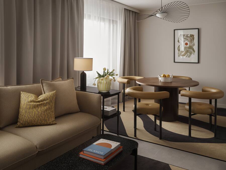 A modern hotel room with a sofa, a coffee table with books, a lamp, a vase with flowers, a dining table with chairs, and a framed picture on the wall.