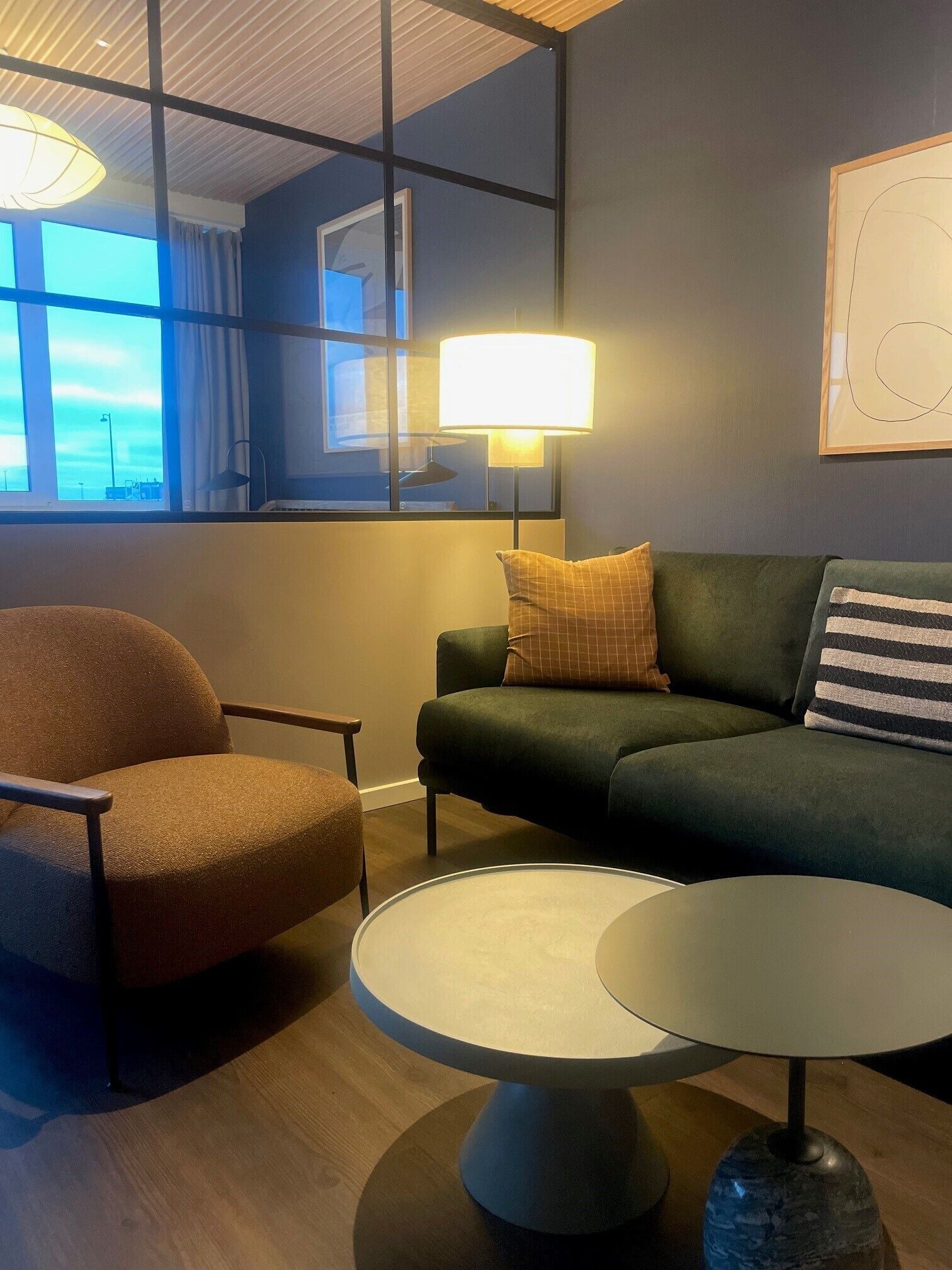 Un moderno salón con un sofá verde, un sillón marrón y una mesa de centro redonda. Se ve un ventanal con cortinas, y una lámpara sobre la mesa.