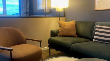 Un moderno salón con un sofá verde, un sillón marrón y una mesa de centro redonda. Se ve un ventanal con cortinas, y una lámpara sobre la mesa.