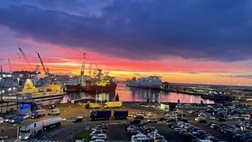 En parkeringsplads med flere biler og et stort skib ved et havneanlæg i solnedgang.