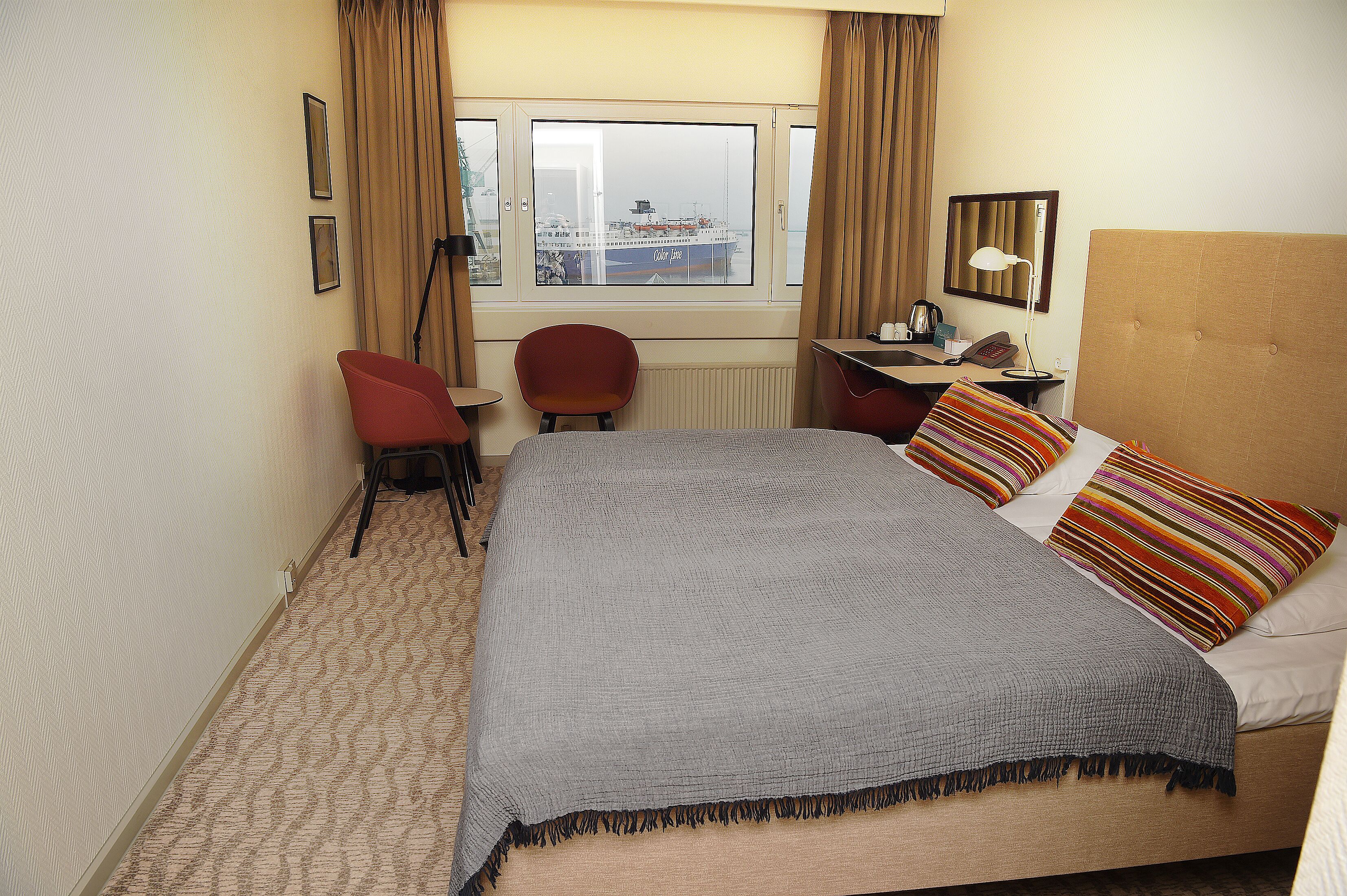Quarto de hotel com uma cama grande, uma escrivaninha, uma cadeira, uma janela com vista para um navio e um abajur.
