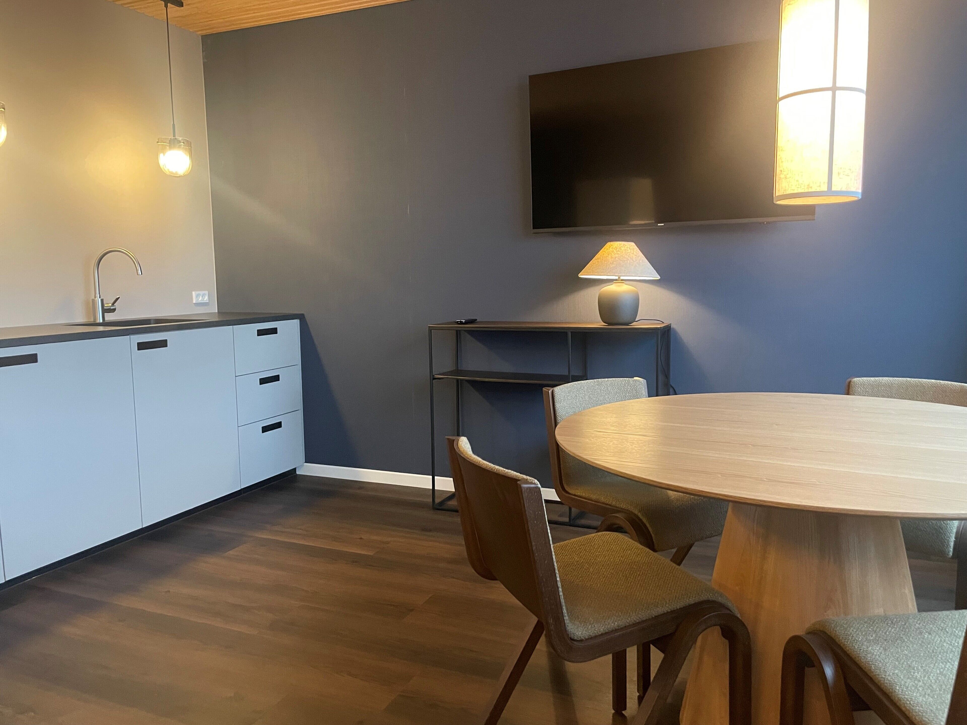 Una cocina moderna con comedor, televisor de pantalla plana en la pared y una lámpara sobre una mesita.