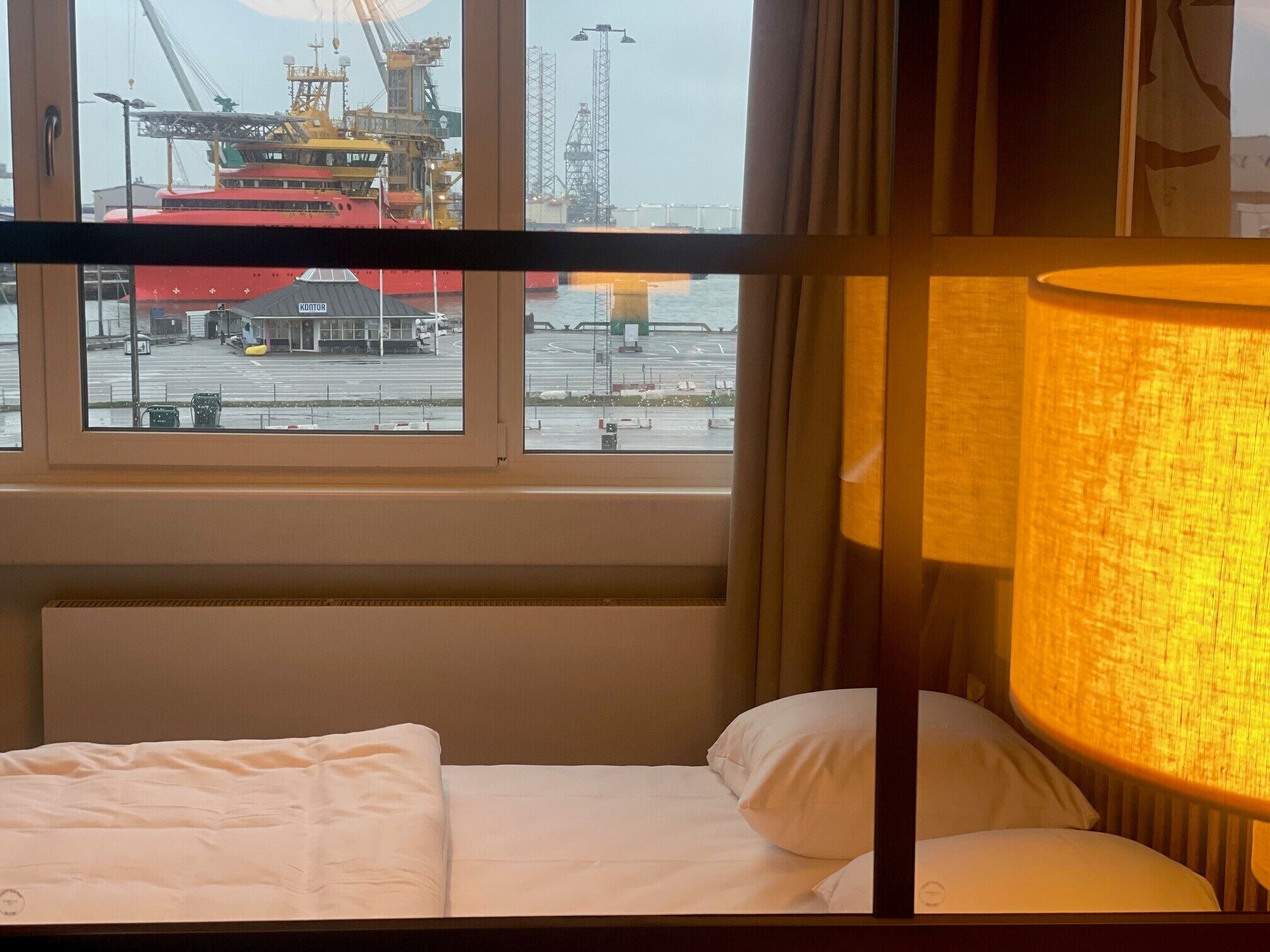 Et hotelværelse med en seng, et vindue med udsigt over en havn og kraner, samt en lampe.