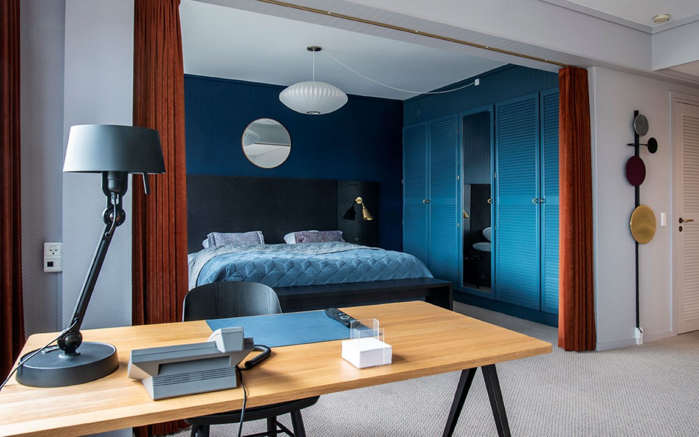 A modern bedroom with a bed, a desk with a lamp, and a wardrobe.