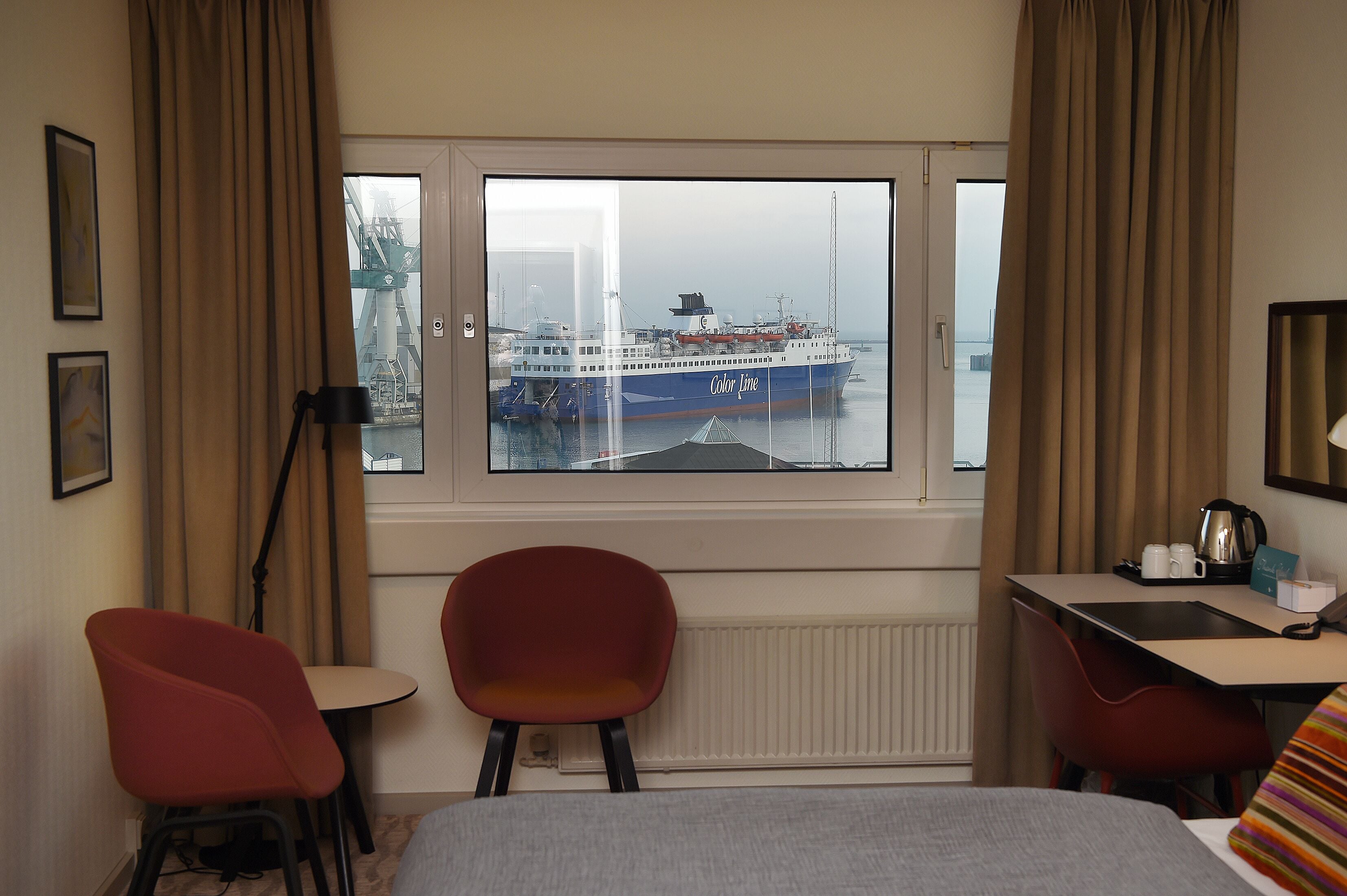 A hotel room with a large window, a bed, a desk, two red chairs, and a radiator.