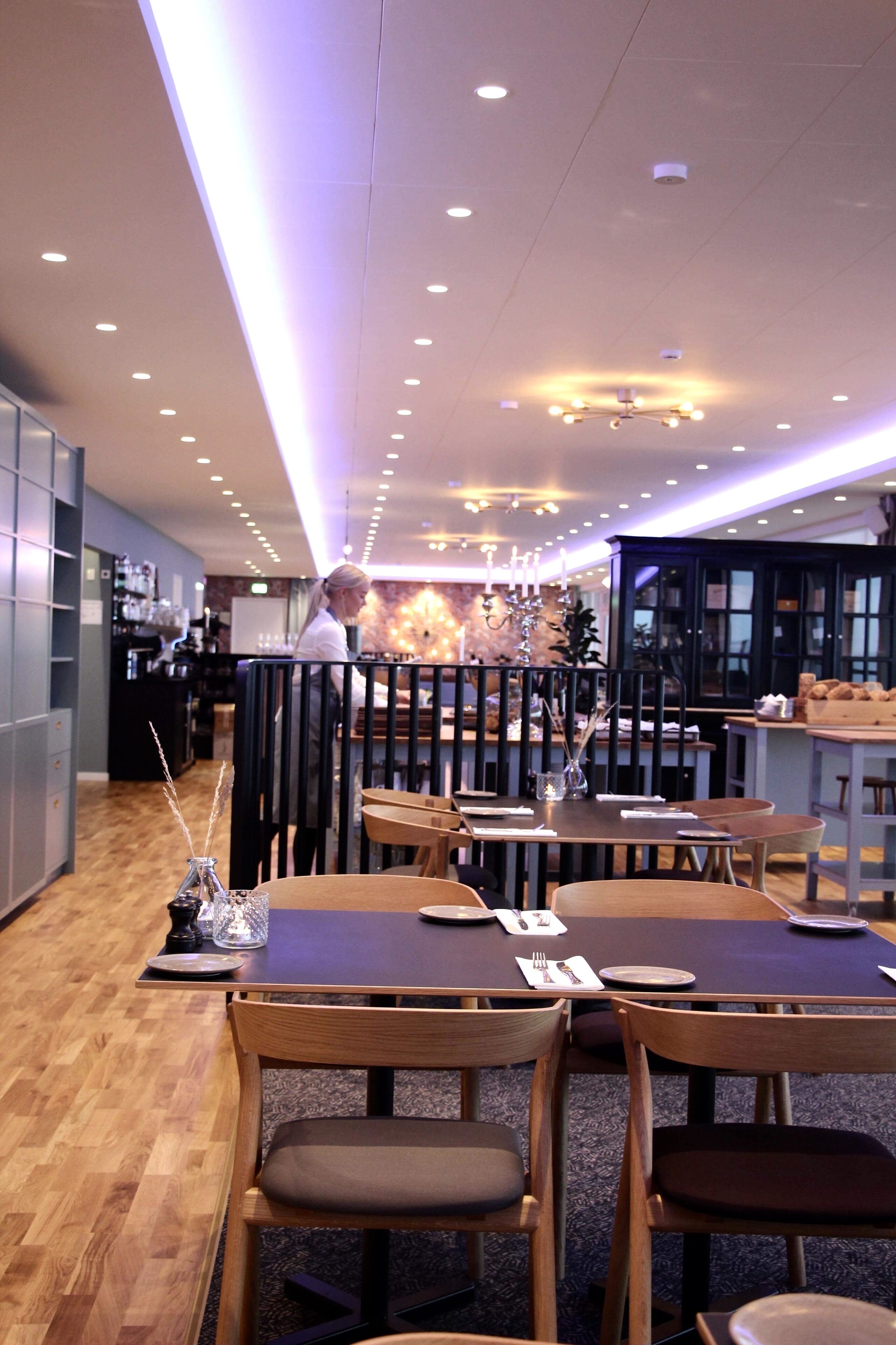 Un intérieur de restaurant moderne avec des tables et des chaises en bois, un bar et un plafond doté d’un éclairage encastré.